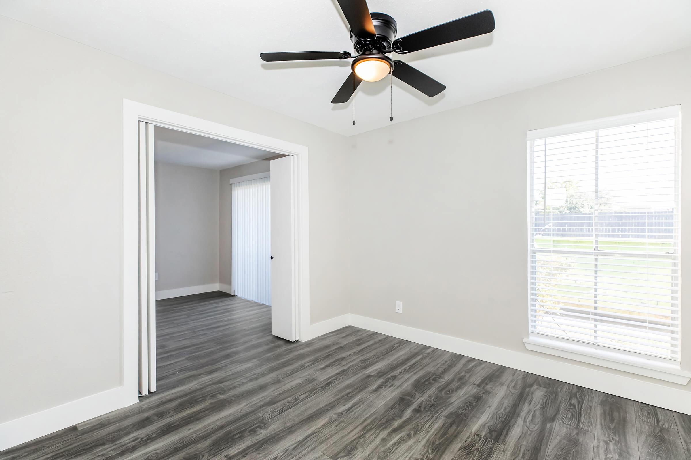 A spacious room with light gray walls and dark wood-like flooring. A black ceiling fan is mounted on the ceiling. To the right, a window with white blinds allows natural light to enter. An open doorway leads to another room with a sliding door and a view of a green outdoor space.