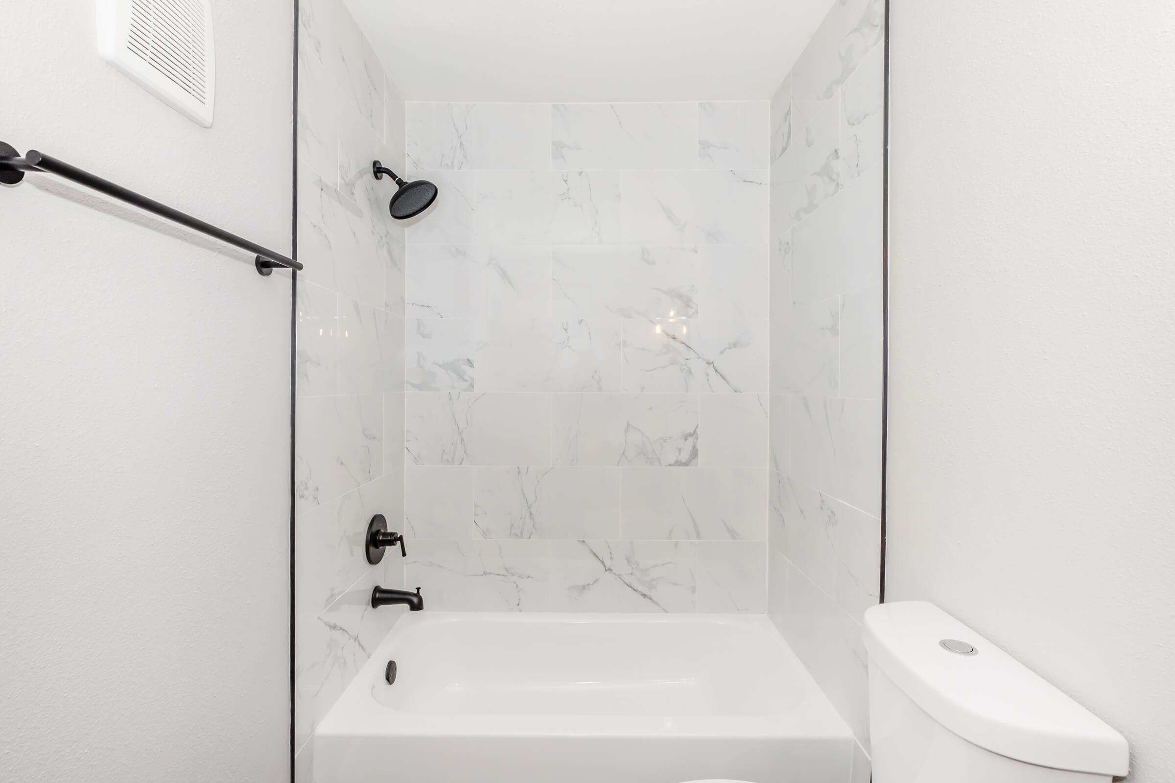 A modern bathroom featuring a white bathtub with a marble-patterned wall. A sleek black showerhead is mounted on the wall, and a black towel rack is visible. The room has a clean and minimalistic design, with a flush toilet positioned next to the bathtub. Bright lighting enhances the overall look.