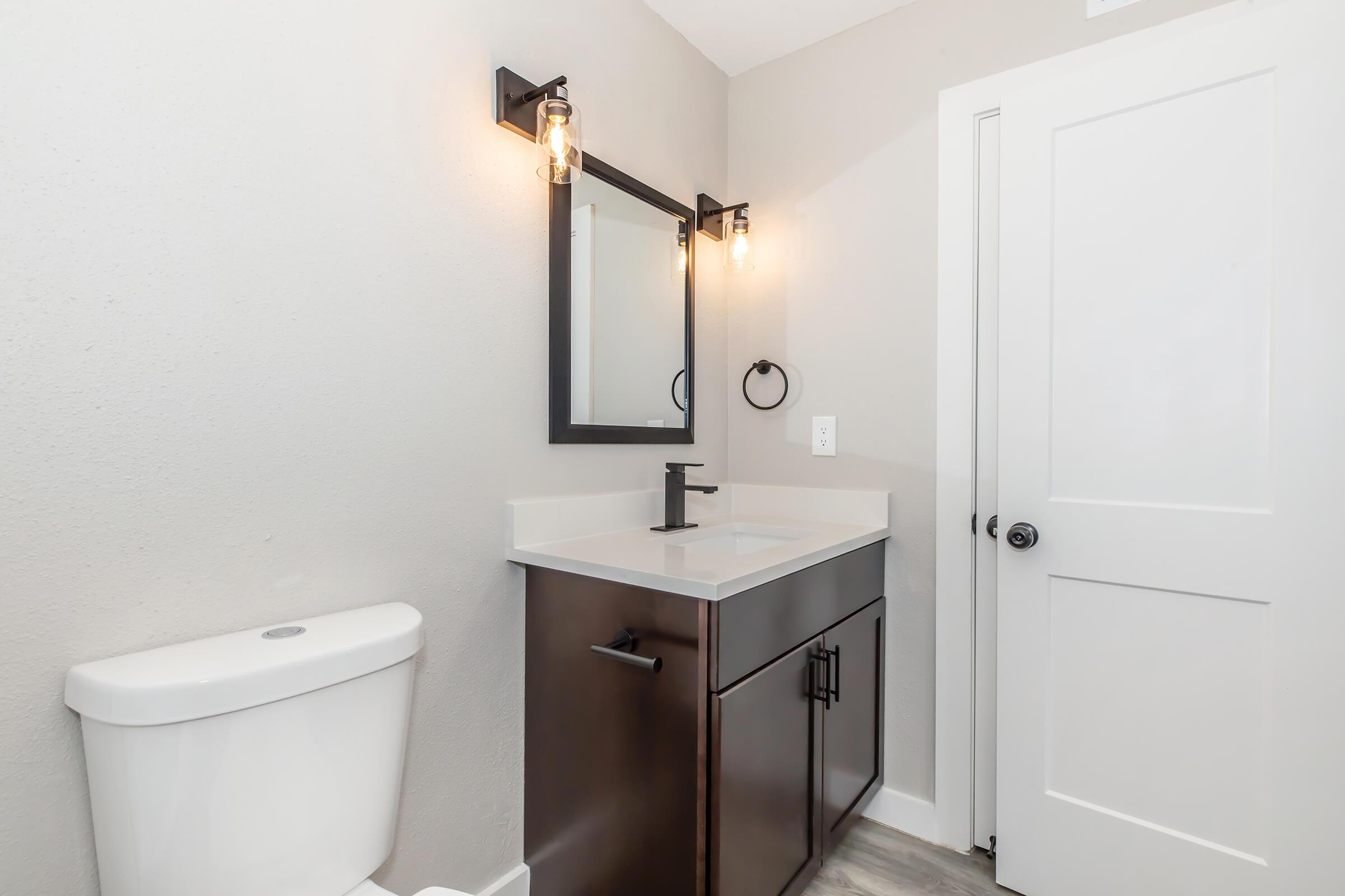 A modern bathroom featuring a sleek dark wood vanity with a white countertop, a rectangular mirror with a black frame, and a stylish light fixture. There’s a white toilet and a door with a simple black handle on the right. The walls are painted in a light gray color, creating a clean and contemporary atmosphere.