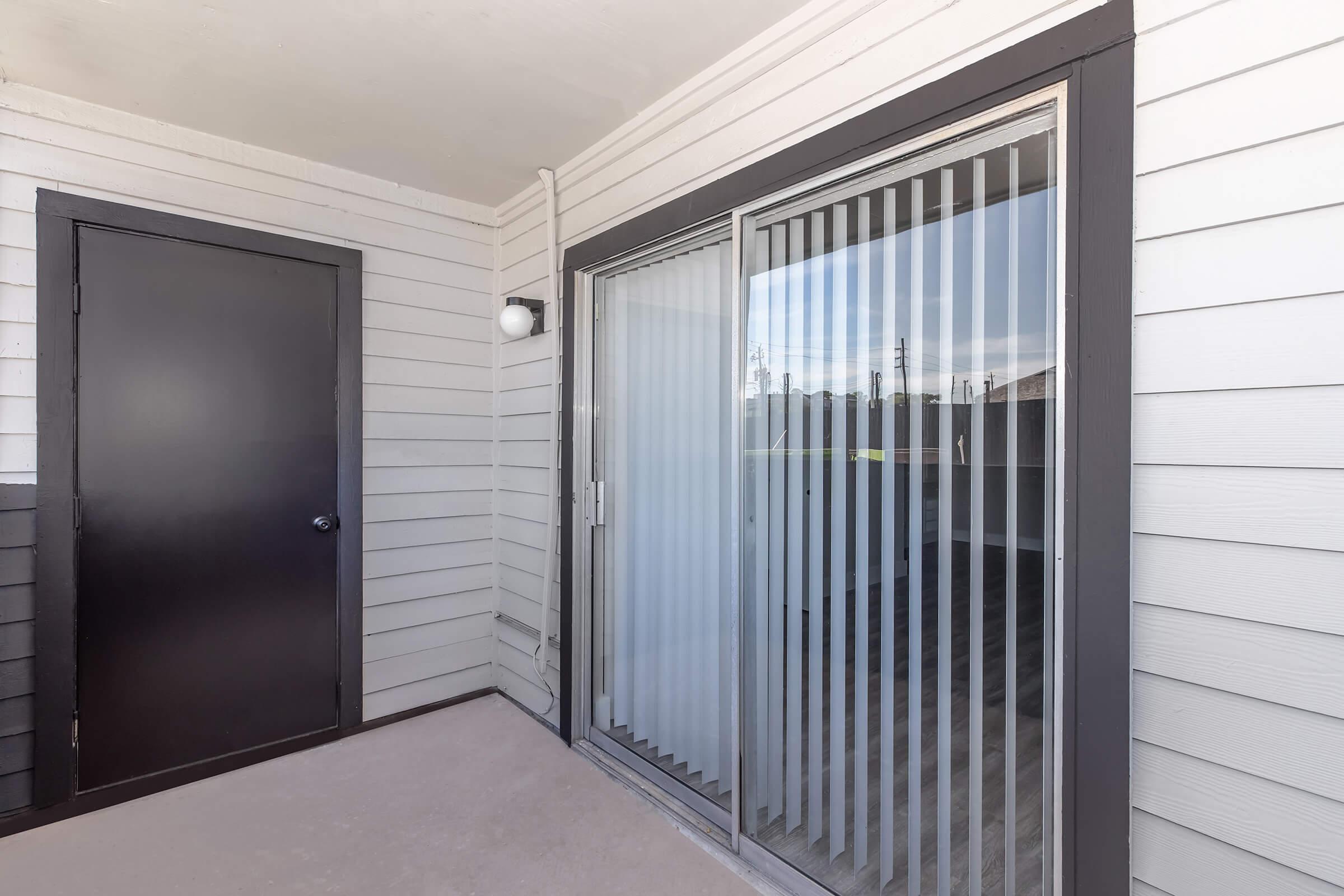 An exterior view of a patio door with vertical blinds, leading to an empty balcony area. There is a closed black door on the left, and the wall is made of light-colored horizontal siding. The patio is well-lit, showing a glimpse of greenery outside.