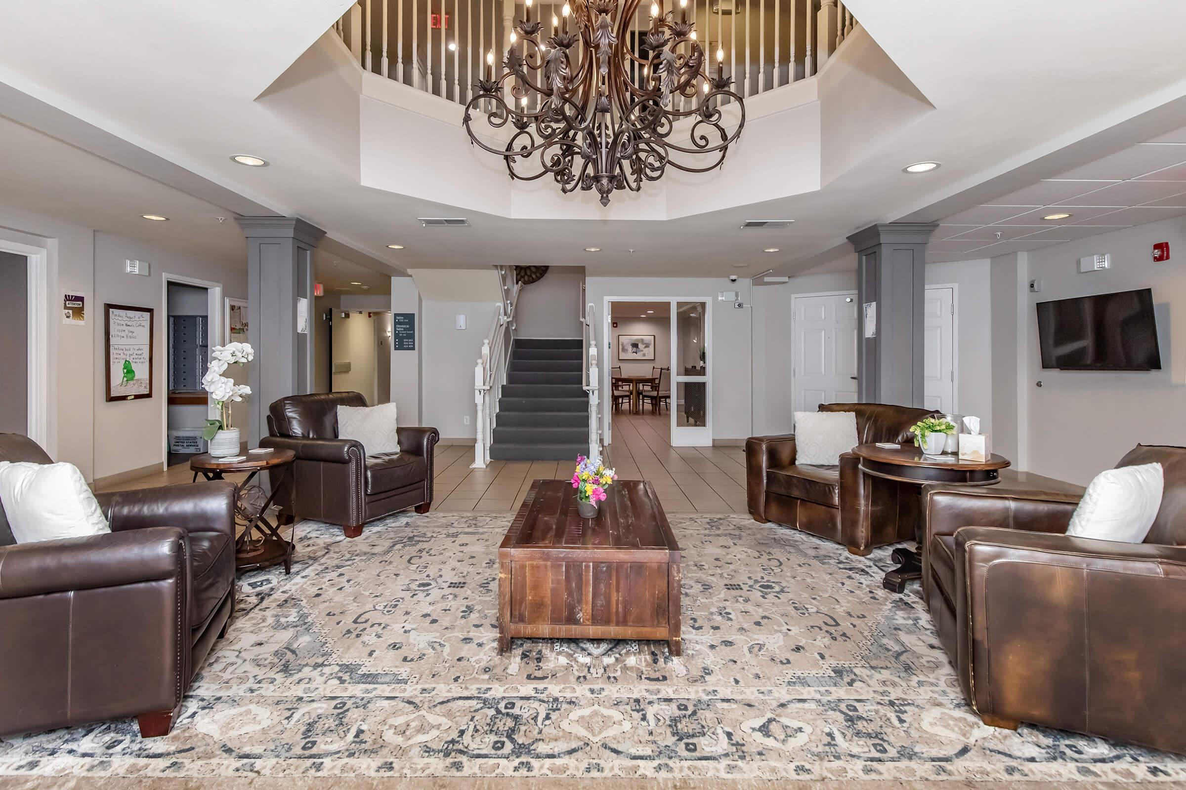 A spacious, well-lit lobby area featuring a large chandelier, comfortable leather chairs, and a wooden coffee table. A floral arrangement sits on the table, and there are staircases leading to further areas. The decor includes elegant light fixtures and a patterned rug, adding to the welcoming atmosphere.