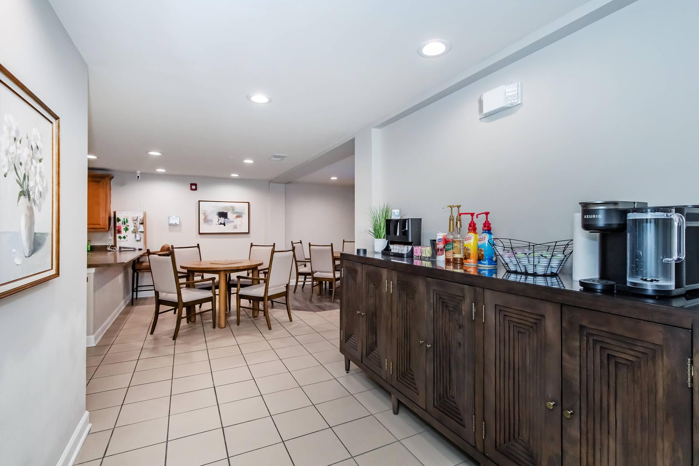 A modern kitchen or dining area featuring a wooden buffet cabinet with various beverages and utensils, a round dining table surrounded by beige chairs, and light gray walls. The space is well-lit, with spotlights and a framed floral artwork on the wall, creating a welcoming atmosphere.