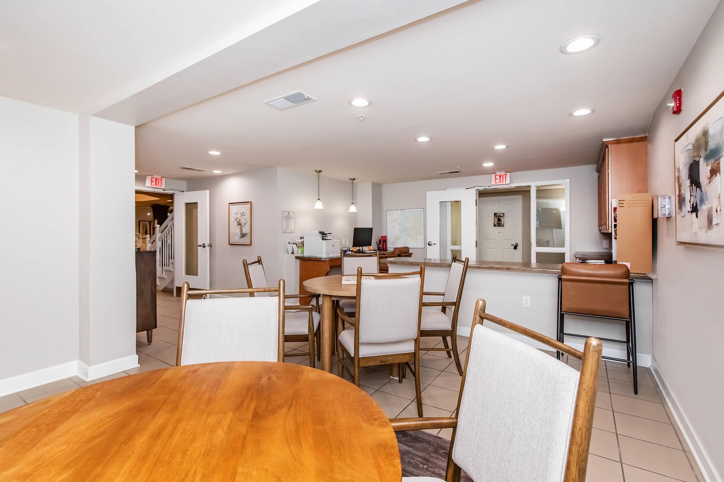 A spacious dining area featuring wooden tables and light-colored chairs. The walls are painted in neutral tones, and there are modern light fixtures. In the background, a reception area is visible with a desk and informational boards. Large windows allow natural light to fill the room.