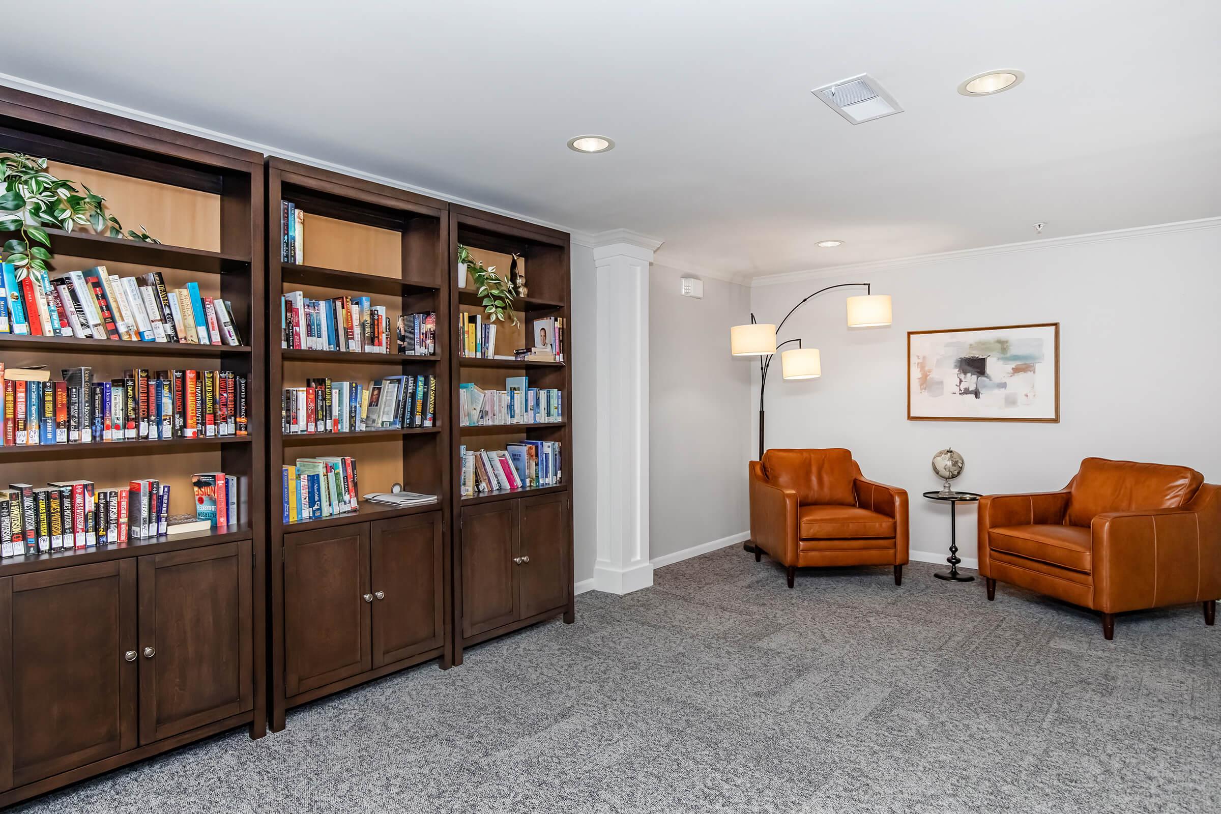 A cozy reading nook featuring two brown leather chairs beside a modern lamp, with a bookshelf filled with various books on the left. The floor is carpeted in gray, and there is a decorative globe on a small table, enhancing the inviting atmosphere of the space.