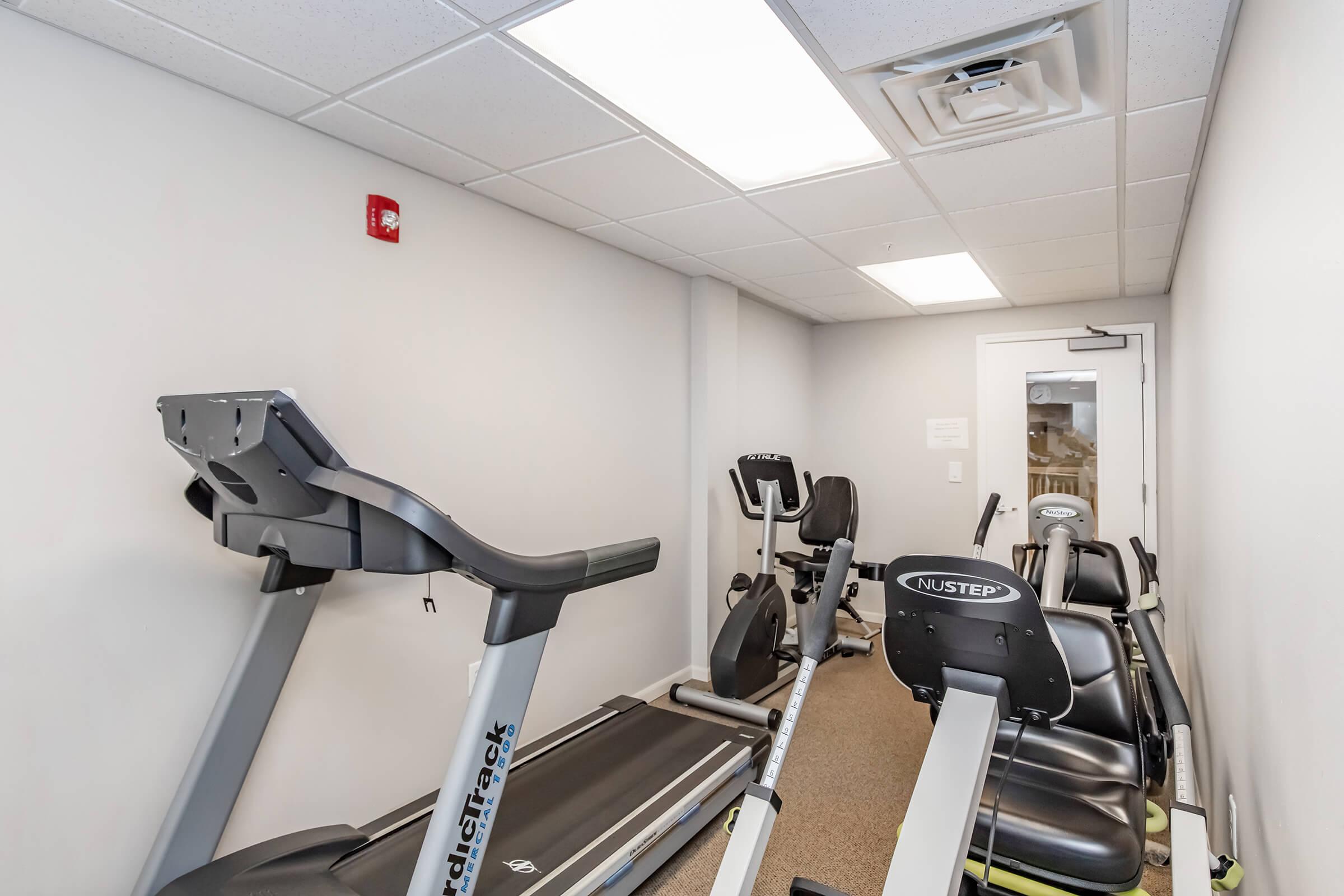 A small fitness room featuring a treadmill, an elliptical machine, and other exercise equipment. The walls are neutral-colored, and the space has overhead lighting. A door leads out to another area, with a clean and organized appearance.