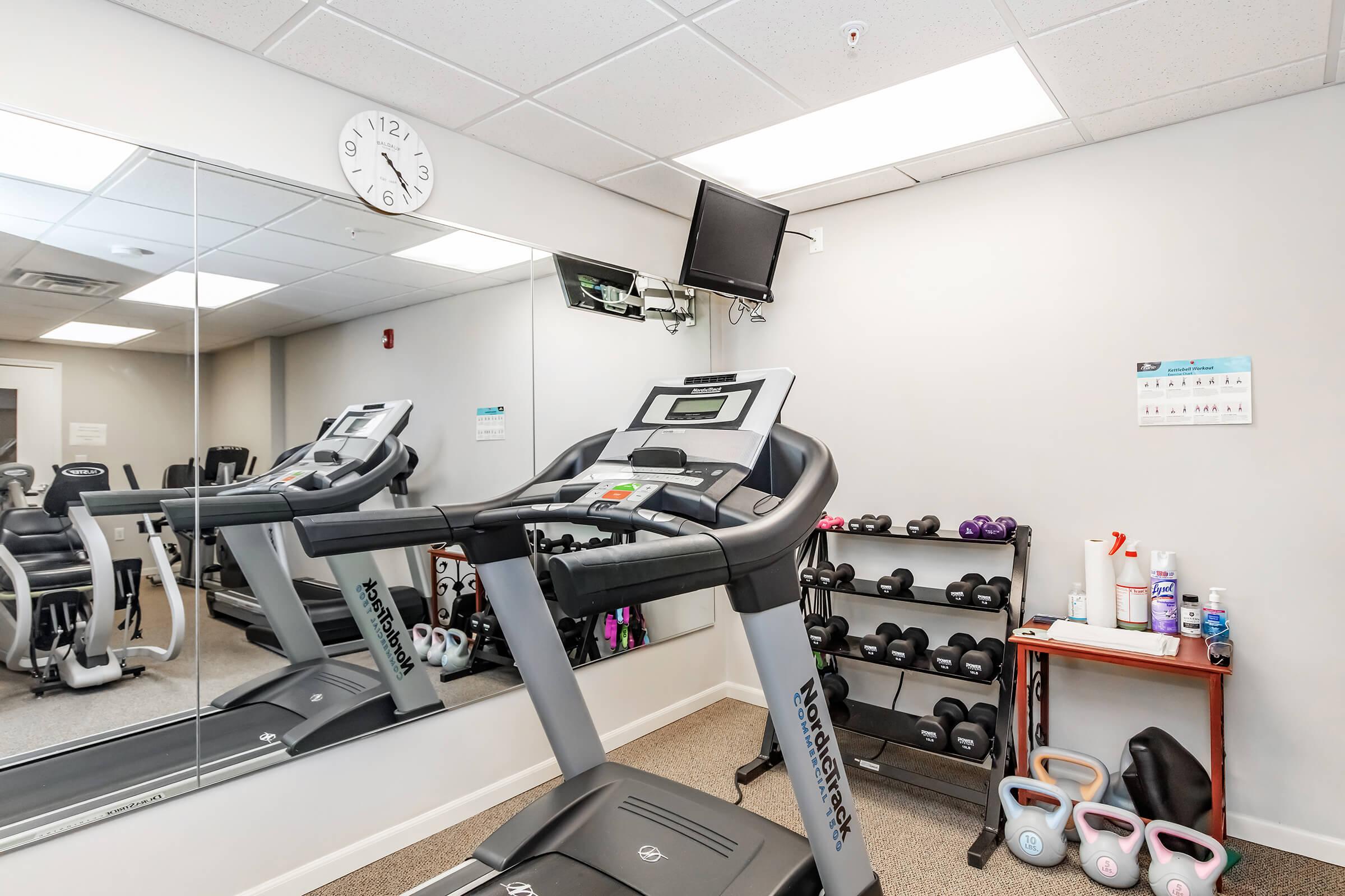 A clean fitness room featuring a treadmill, exercise equipment, a wall mirror, and a wall-mounted TV. Weights are neatly arranged on a rack, and a calendar is visible on the wall. The floor is carpeted, creating a welcoming exercise environment.