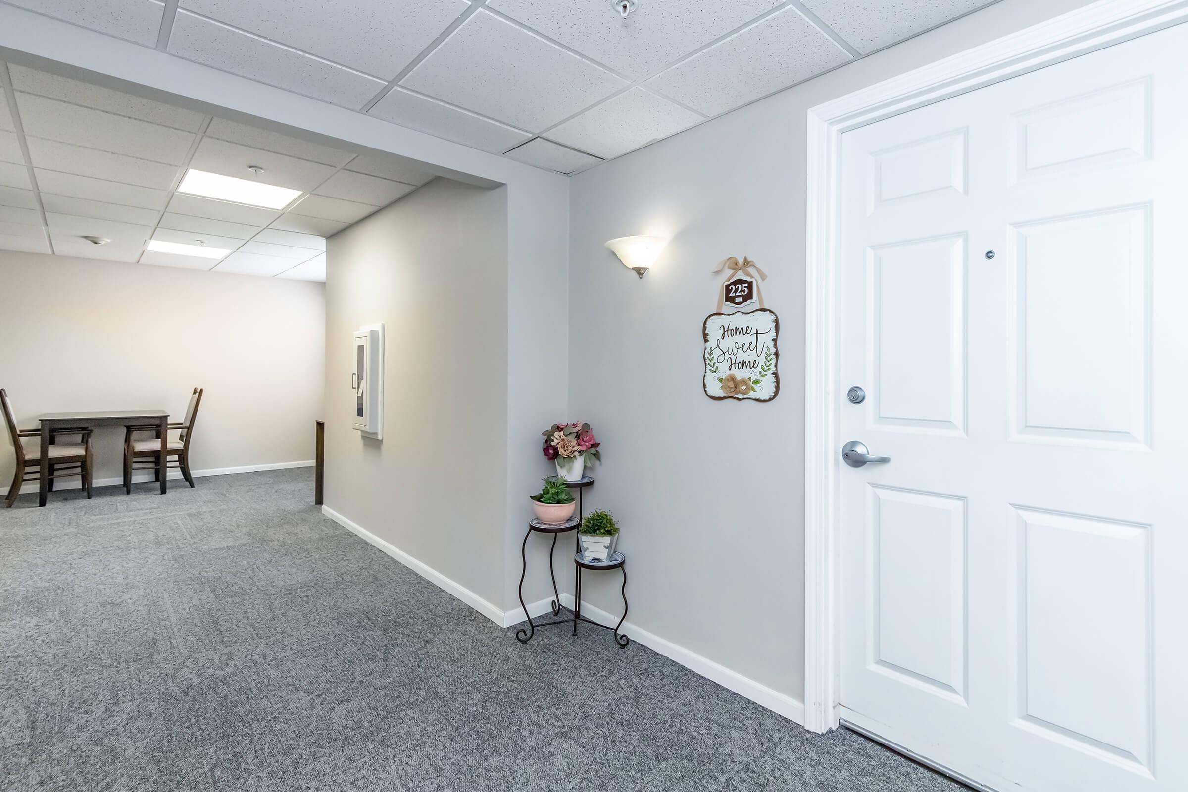 A well-lit hallway with gray carpet leading to a closed white door marked "235." To the side, a small decorative plant stand holds flowers. The walls are painted in a light color, and there’s a dining area visible in the background with a table and chairs.
