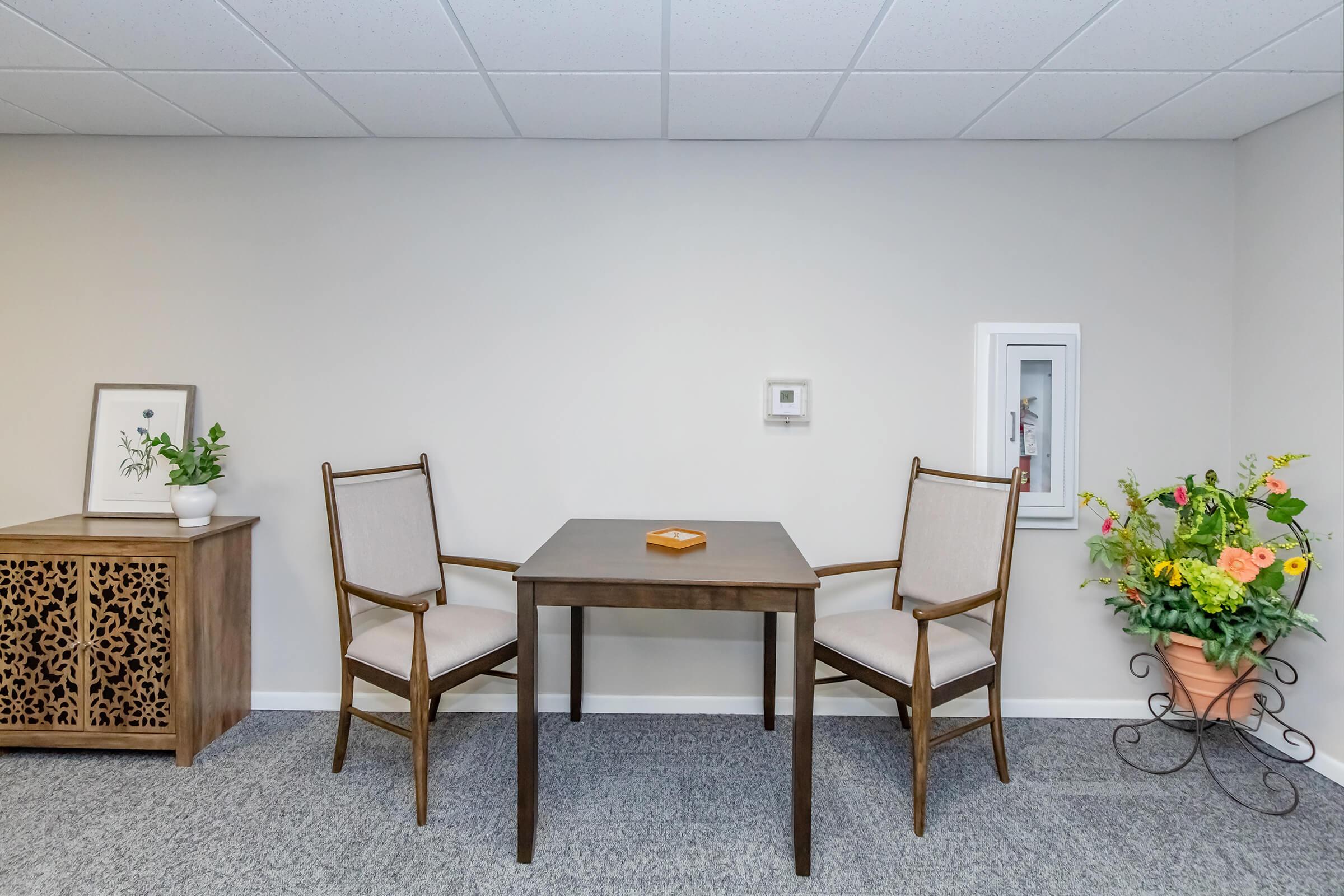 A simple interior scene featuring a small wooden table with two matching chairs. To the left, there's a decorative cabinet with an intricate design, and on the right, a wall-mounted fire safety box. A vibrant flower arrangement adds a touch of color, enhancing the warm, inviting atmosphere of the space.