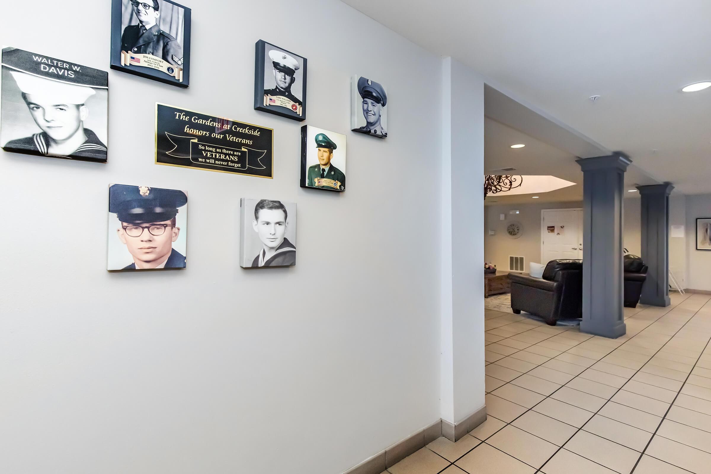 A hallway featuring framed portraits of veterans on the wall, with a plaque dedicated to honoring their service. The area is well-lit, showcasing the images and a comfortable seating area in the background.
