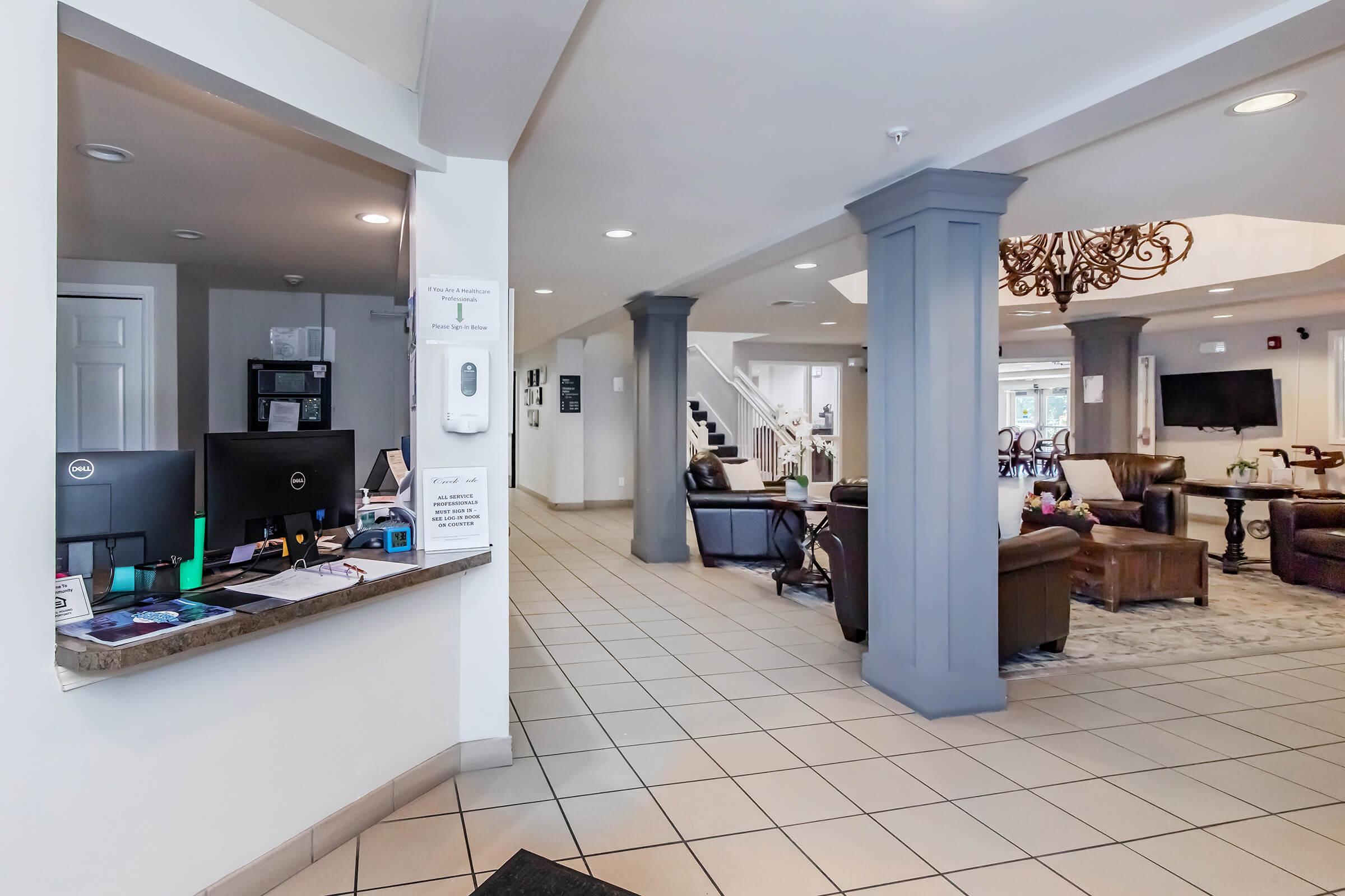 Interior view of a lobby area featuring a reception desk on the left with computers and office supplies. The space has a modern design with a chandelier hanging from the ceiling, comfortable seating areas, and a staircase in the background. The floor is tiled and well-lit, creating an inviting atmosphere.