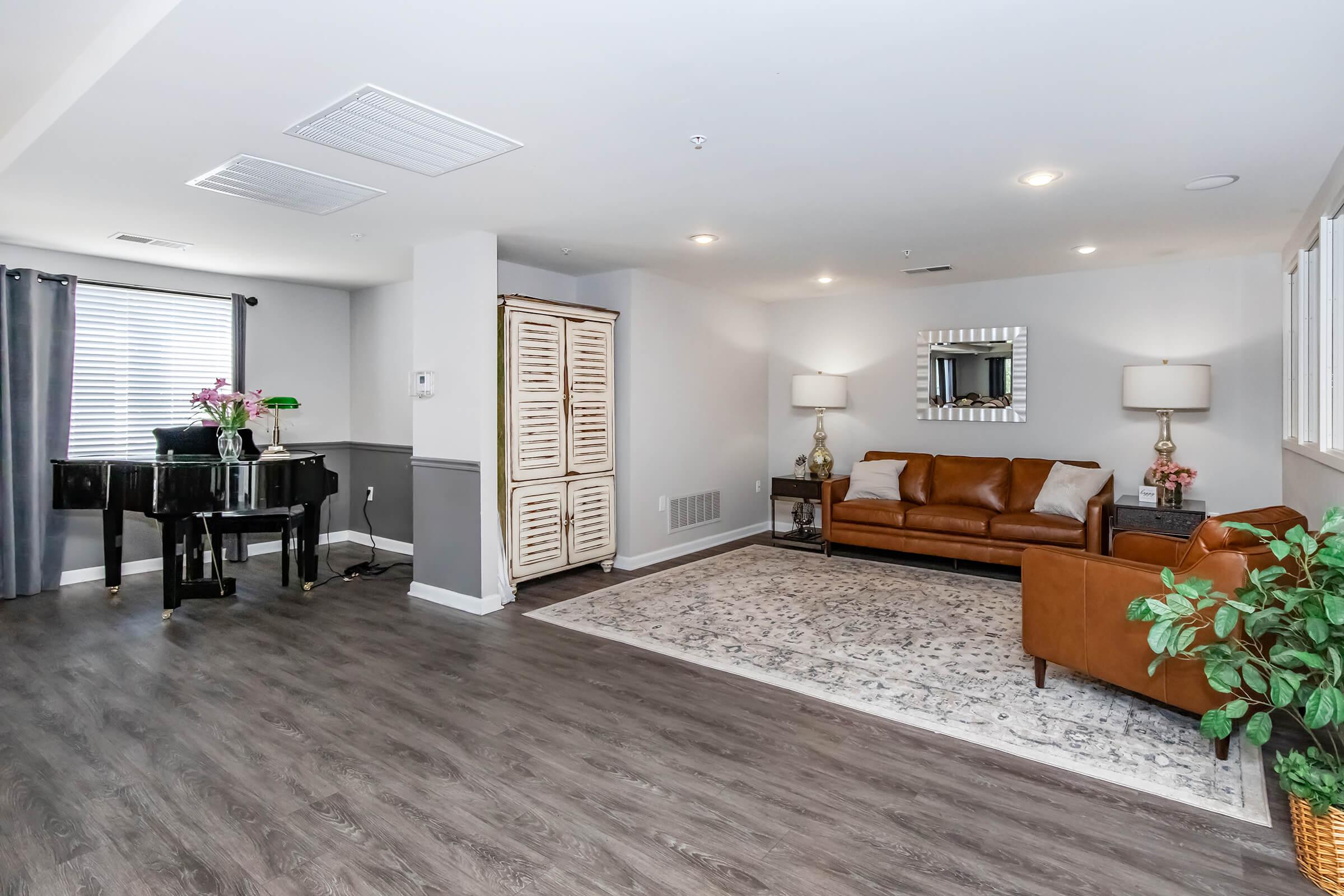 A spacious modern living area featuring a brown leather sofa, a glass-top coffee table, and a piano. The room has gray walls, hardwood flooring, and a decorative area rug. Natural light filters through window treatments, while a mirror and decorative plants add visual interest to the space.