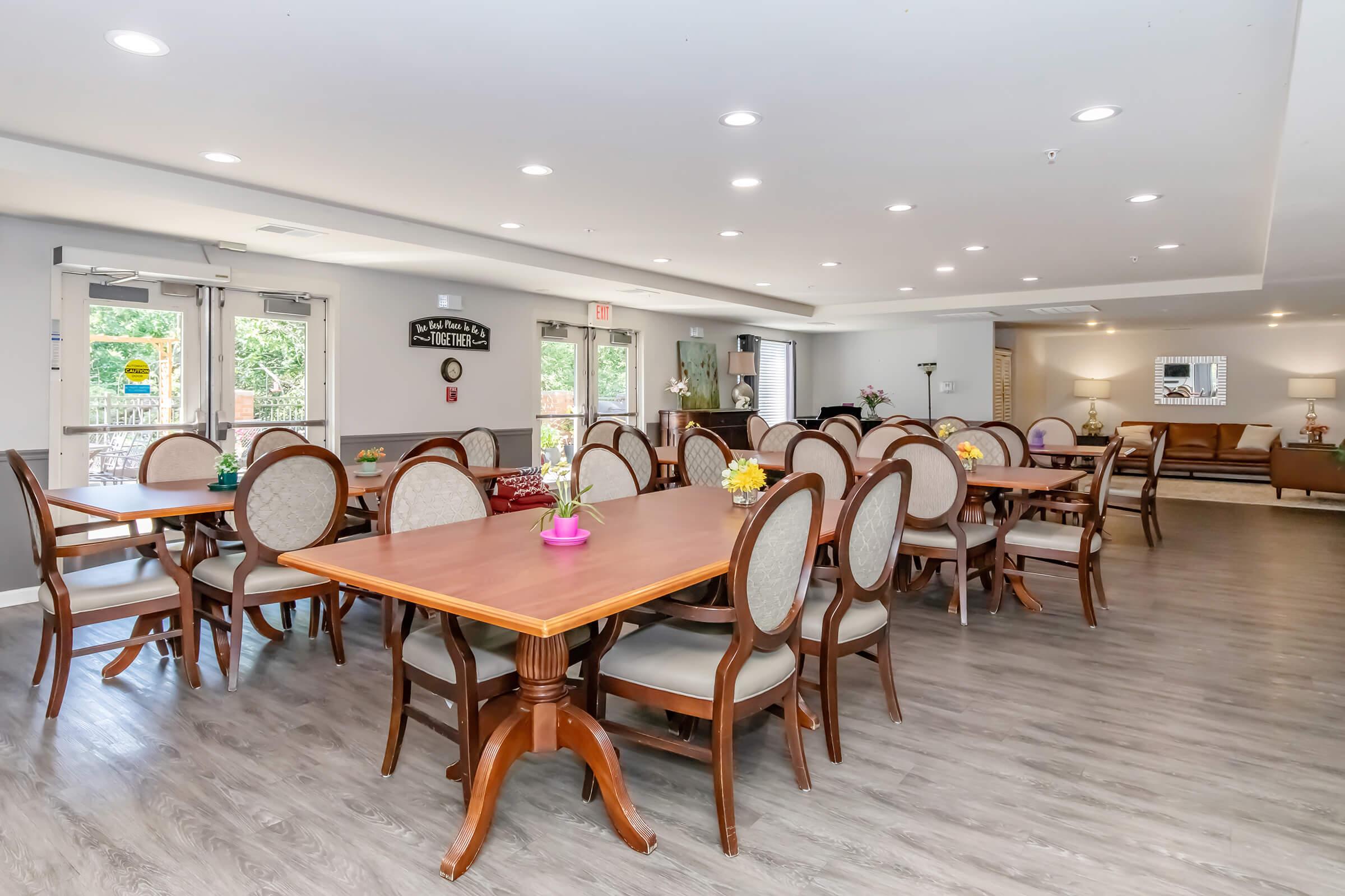 A bright and spacious dining area featuring multiple wooden tables with elegant chairs, neatly arranged in a well-lit room. Decor elements include small flower vases on the tables and large windows allowing natural light. A cozy seating area is visible in the background, adding to the inviting atmosphere.