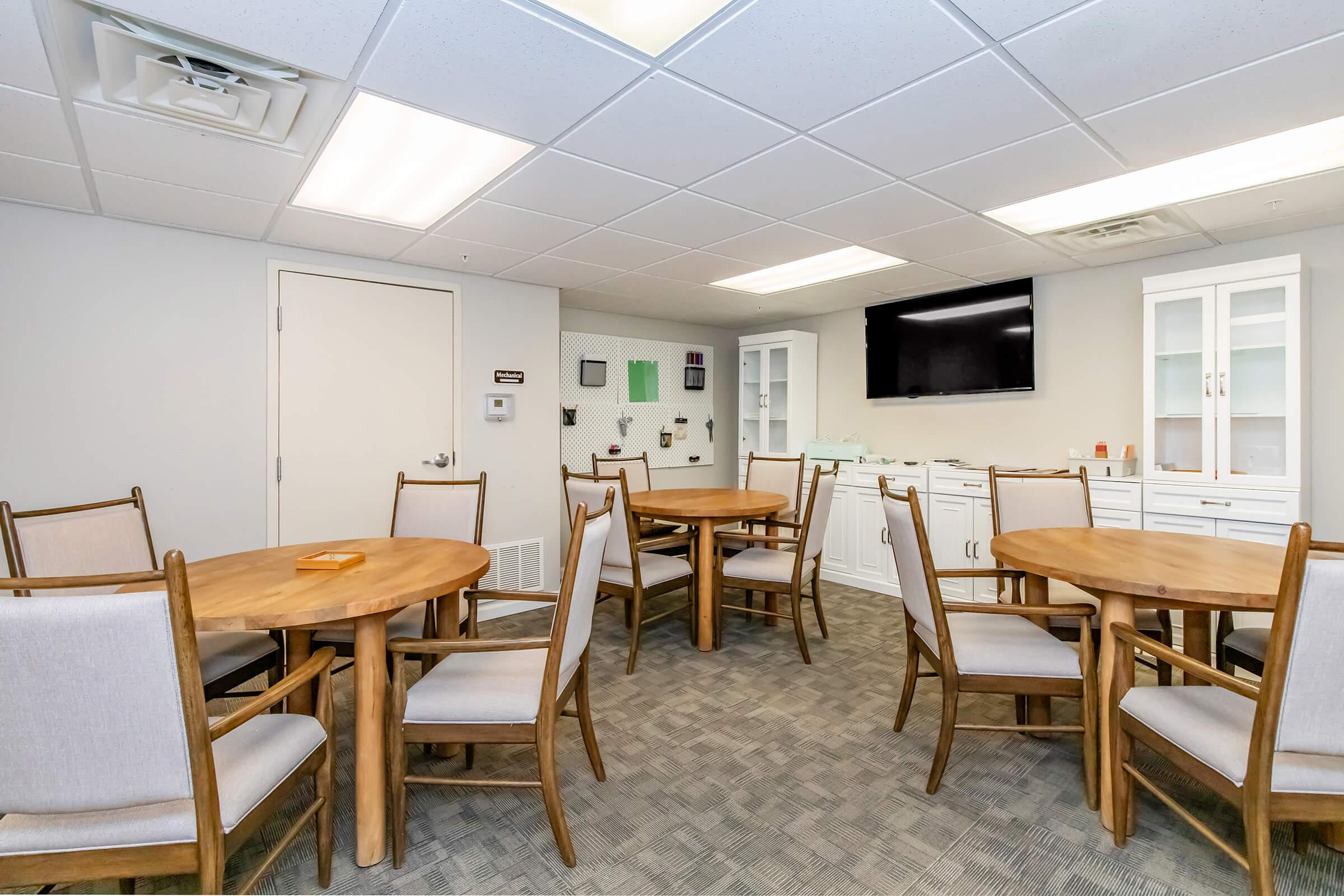 A multiple seating arrangement in a meeting room with several round wooden tables and chairs. The room features light-colored walls and a flat-screen TV mounted on one wall. There are cabinets and a whiteboard visible in the background, creating a professional atmosphere for discussions or presentations.