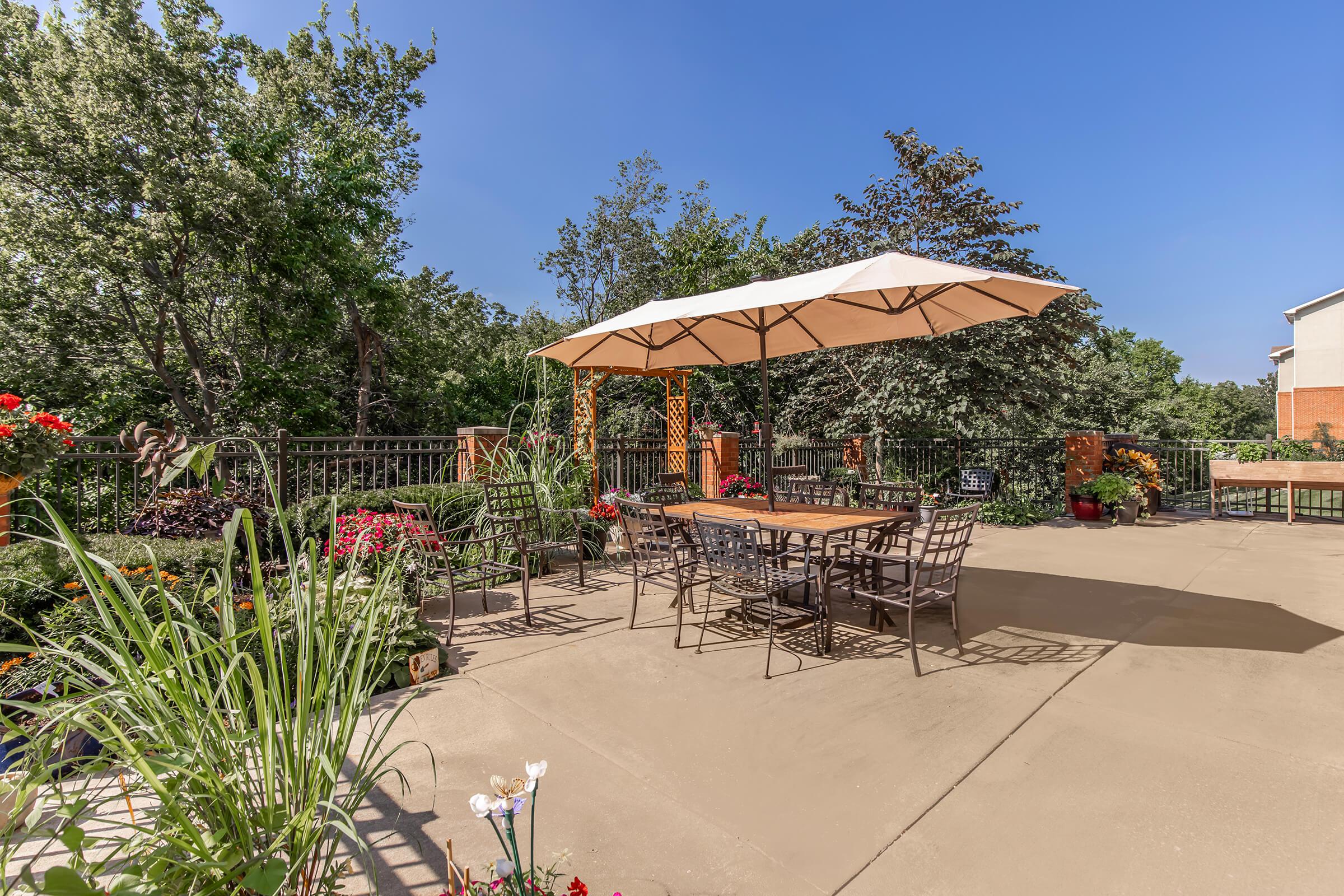 A spacious outdoor patio featuring a large umbrella over a dining table surrounded by metal chairs. The area is adorned with vibrant flower beds and lush greenery, with trees in the background and a clear blue sky. The setting suggests a relaxing environment for outdoor gatherings.
