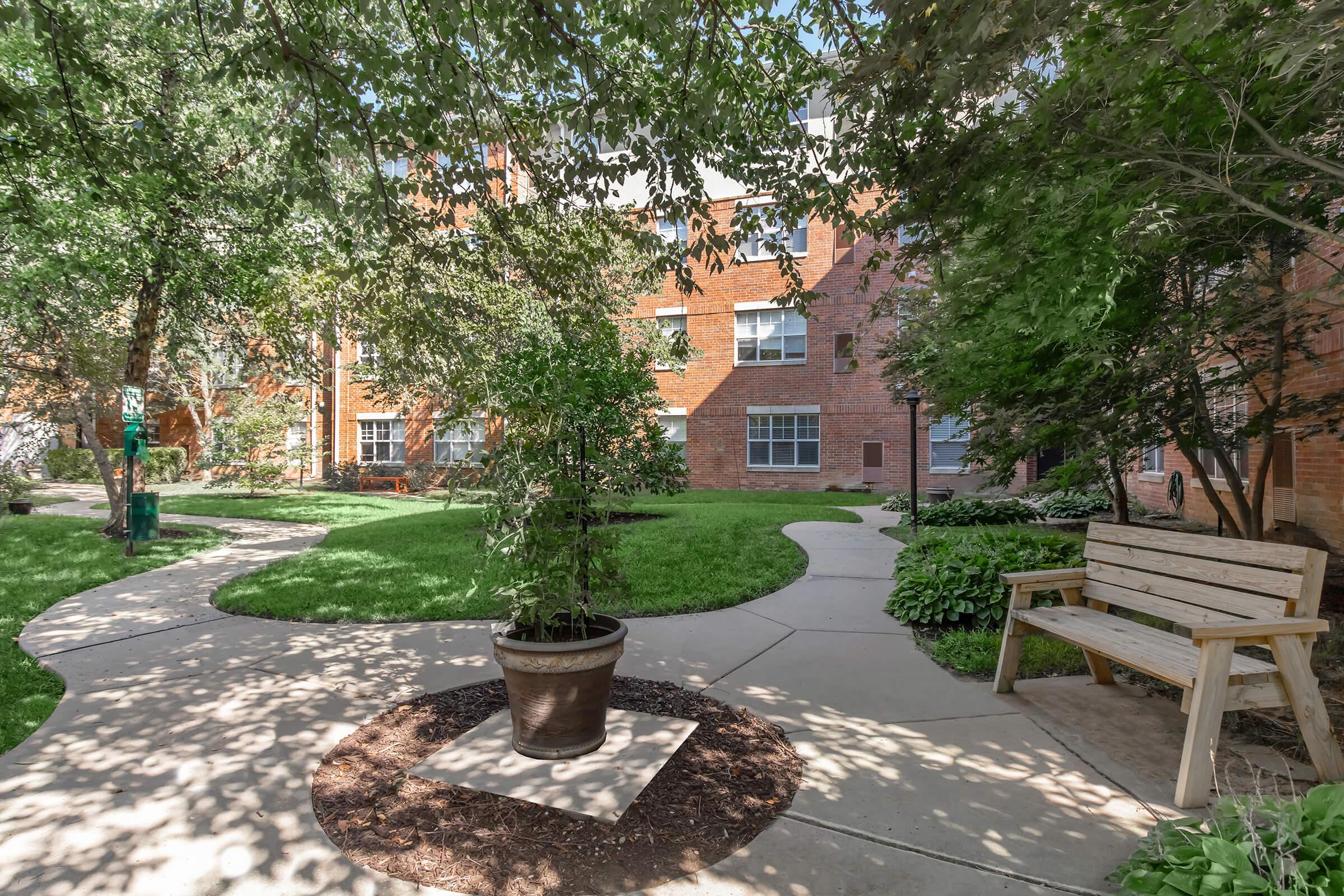 A landscaped courtyard featuring green grass, trees, and shrubs. Curved concrete pathways lead through the area, with a wooden bench near a potted plant. The background shows brick buildings, creating a serene and inviting atmosphere.