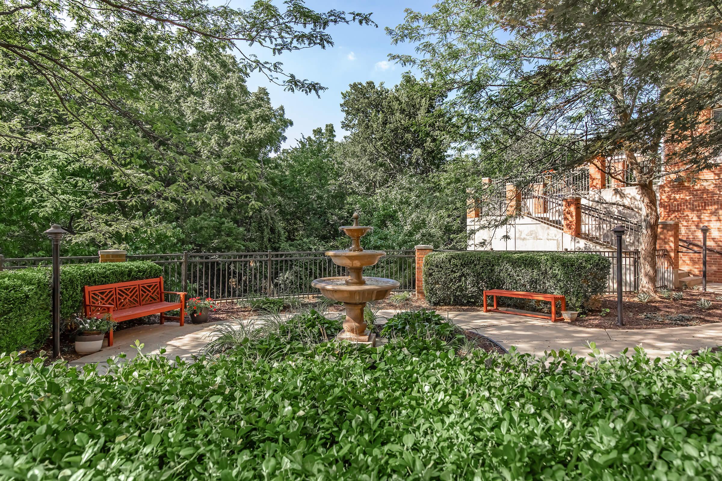 A picturesque garden scene featuring a stone fountain at the center, surrounded by lush greenery and neatly trimmed hedges. There are two red benches positioned along a paved walkway, bordered by a black wrought-iron fence and trees in the background, creating a peaceful and inviting outdoor atmosphere.