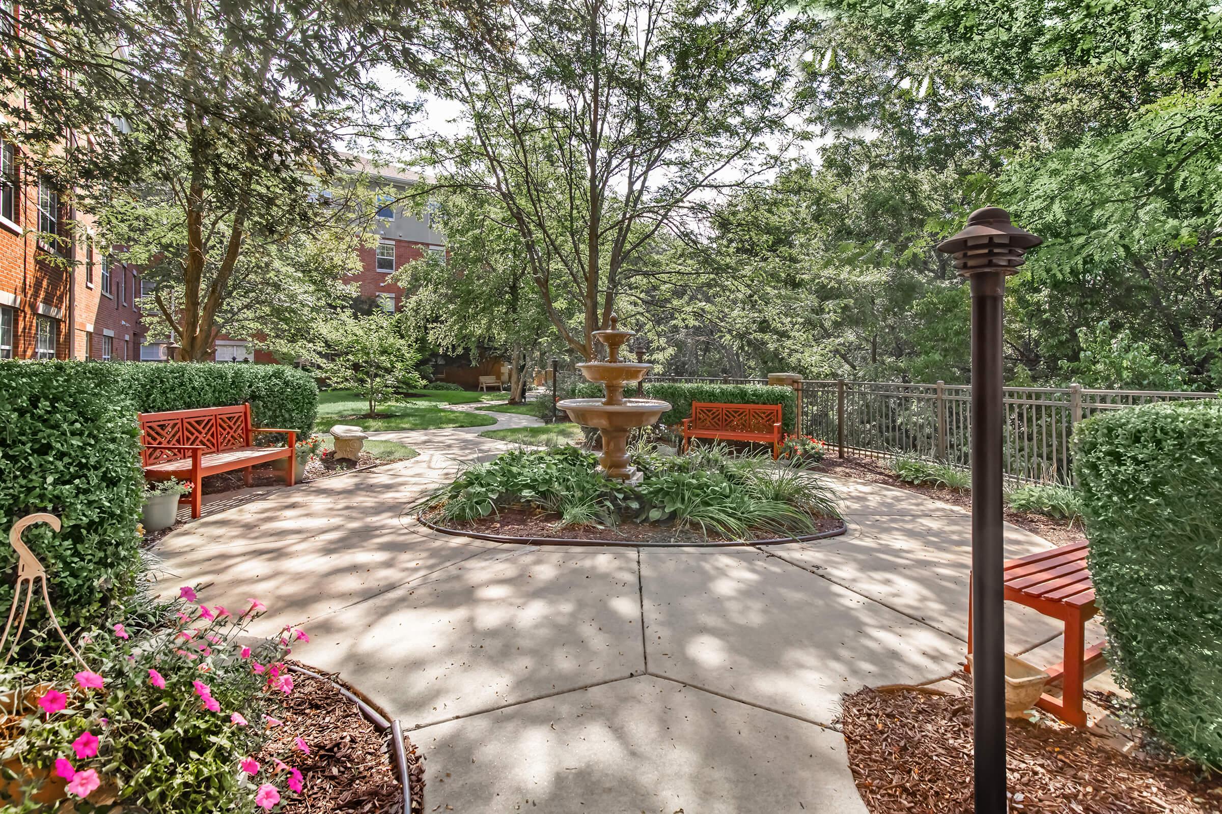 A tranquil garden featuring a three-tiered fountain surrounded by lush greenery, flower beds, and well-maintained pathways. Two wooden benches provide seating, inviting relaxation in this serene outdoor space. The scene is bathed in natural sunlight, enhancing the peaceful ambiance.