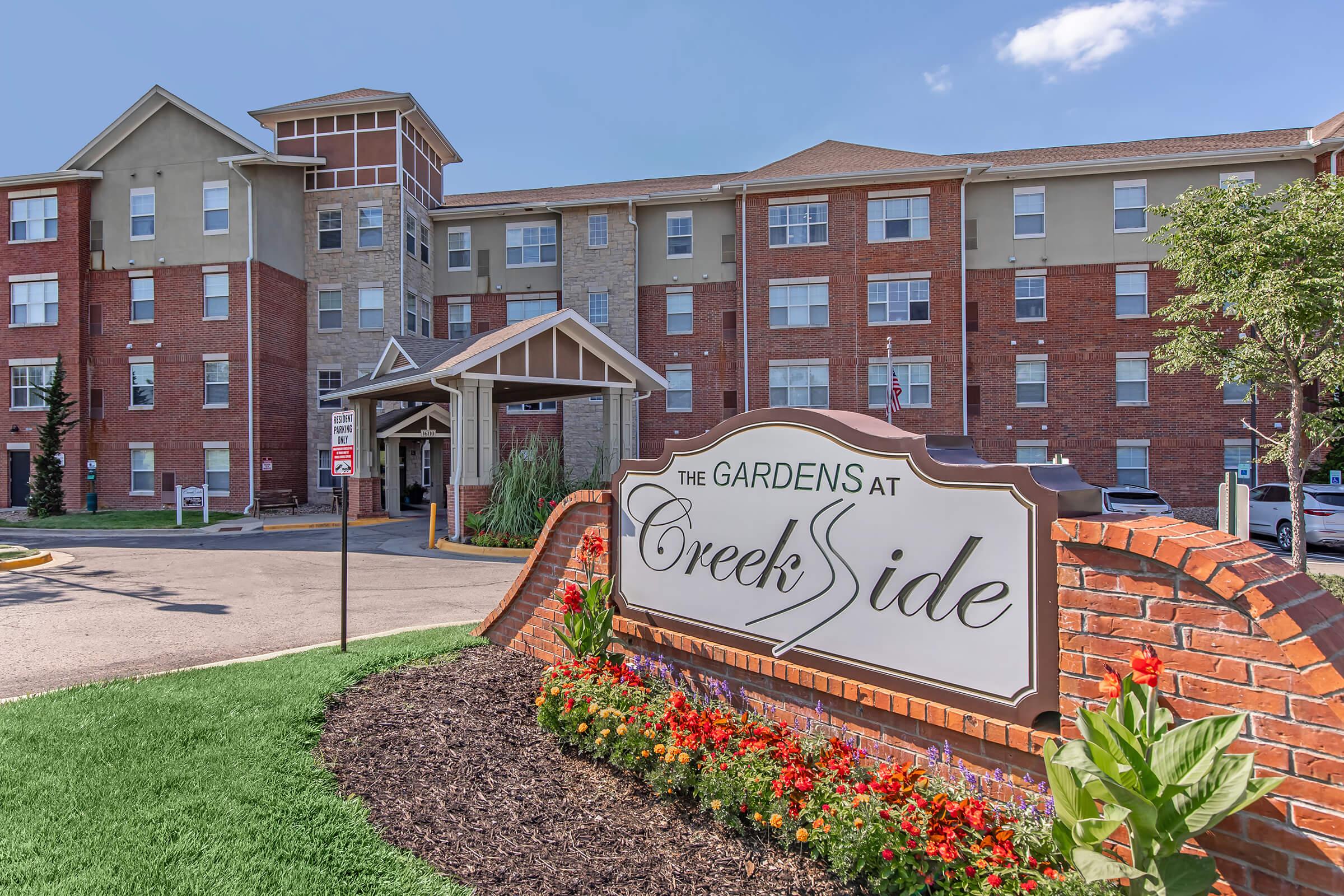 A brick sign displaying "The Gardens at Creekside" in front of a multi-story residential building. The building features a mix of brick and light-colored siding. There are flower beds with colorful blooms in the foreground and a clear blue sky overhead, creating a welcoming atmosphere.