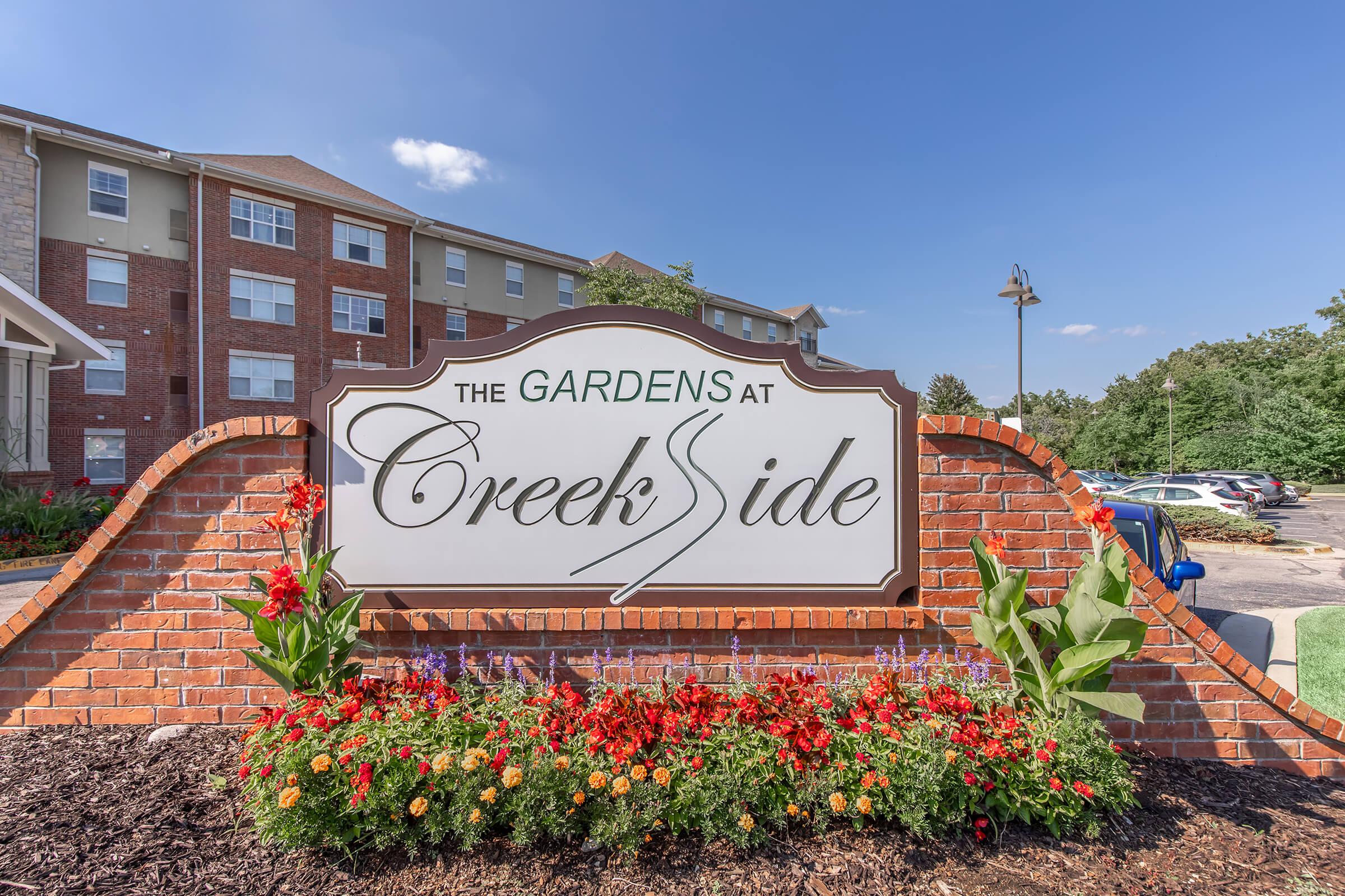 Signage for "The Gardens at CreekSide," featuring a brick structure with colorful flowers and green plants in front. The background includes a modern building under a clear blue sky, indicating a welcoming community atmosphere.