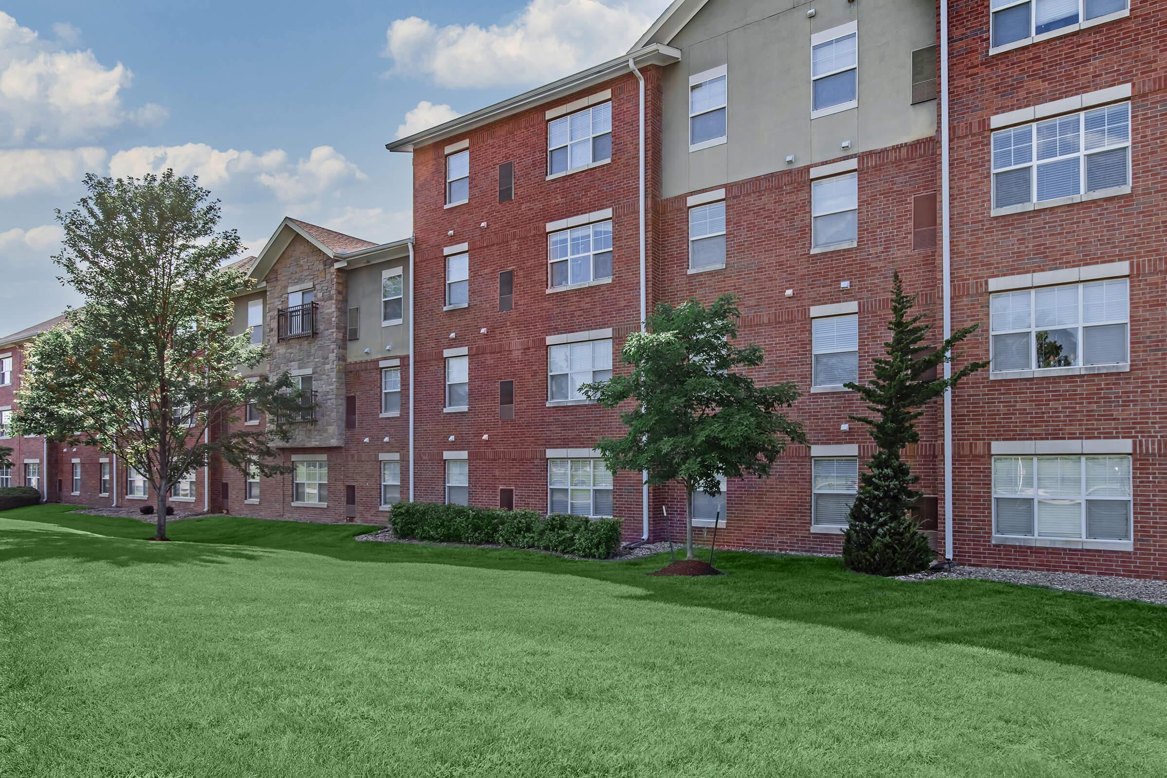 A brick apartment building with multiple windows, surrounded by green grass and small trees. The sky is partly cloudy, adding to the serene atmosphere of the residential area.
