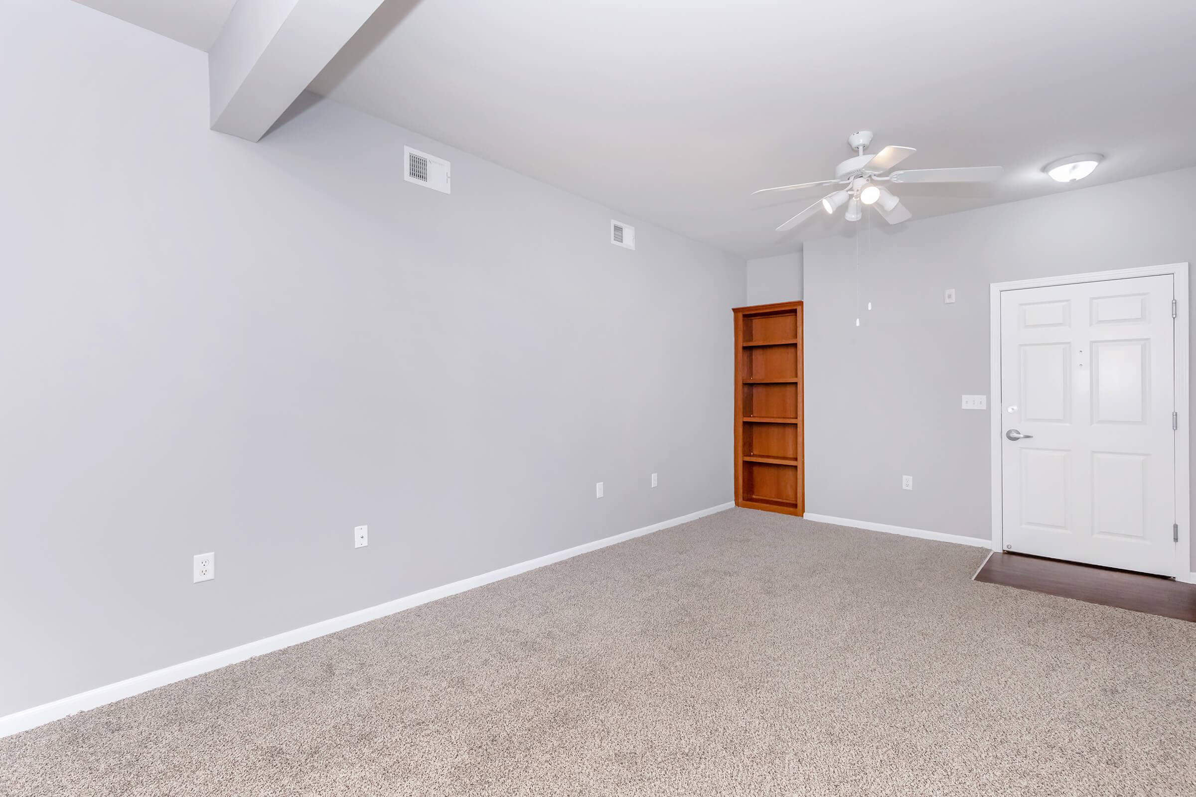 Empty room with gray walls and carpet flooring, featuring a ceiling fan. There is a small built-in bookshelf against one wall and a front door with a white frame and doorknob. The space is well-lit, presenting a clean and modern appearance.