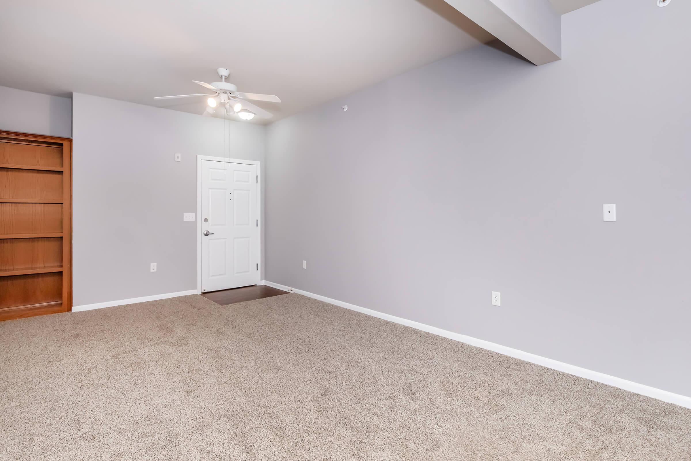 Empty interior space featuring light gray walls, carpeted floor, and a ceiling fan. To the left, there is a wooden bookshelf with an open area on the right that includes a door leading outside. The room is well-lit with natural light, creating a spacious and inviting atmosphere.