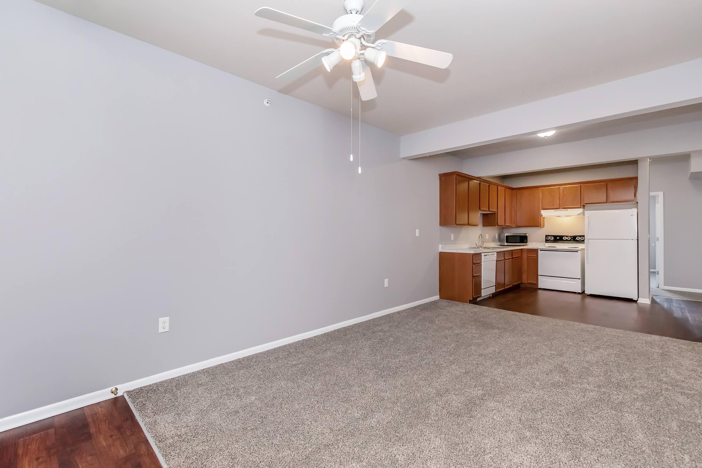 Spacious living area with light gray walls and a ceiling fan. Plush carpet covers the floor, leading to an open kitchen with wooden cabinets, a white refrigerator, and a stove. The layout creates a bright and inviting atmosphere, perfect for comfortable living.