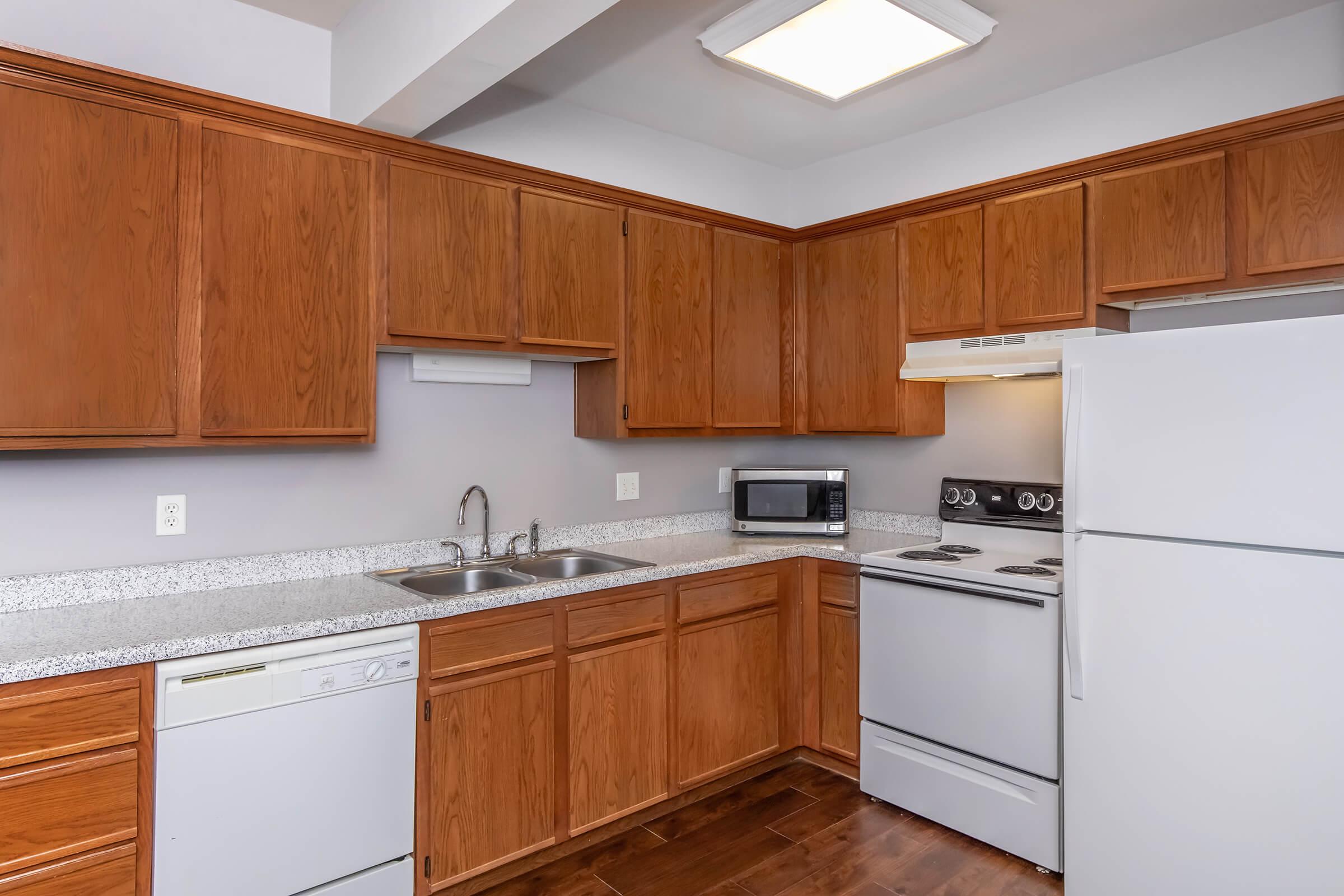 Modern kitchen featuring wooden cabinets, a gray countertop, a double sink, a stove, and a refrigerator. Includes a built-in microwave and a dishwasher, with hardwood flooring and neutral wall colors, creating a clean and inviting cooking space.