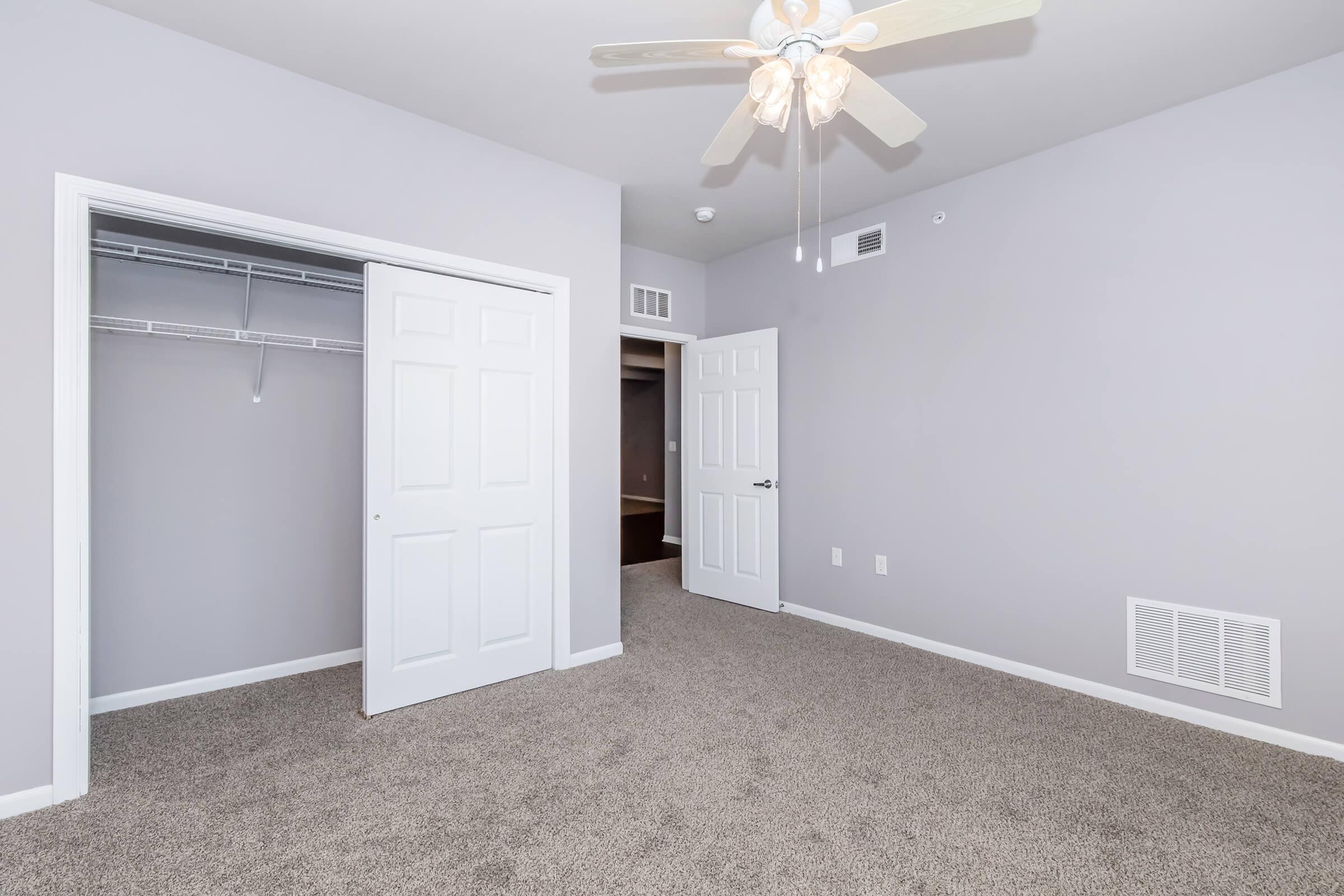 A spacious, light gray bedroom featuring a ceiling fan, soft carpet, and two doors leading to closets. The walls are neutral, creating a calm atmosphere. Natural light may enter from a window not shown in the image.