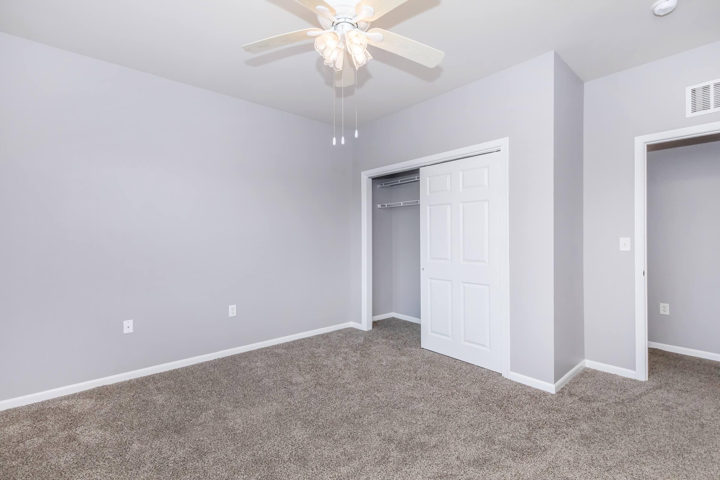 A spacious, empty bedroom featuring light gray walls, carpeted flooring, and a ceiling fan. There is a closet with sliding doors on one side, and another door leading to an adjoining room. The overall atmosphere is bright and neutral, suitable for personalization.