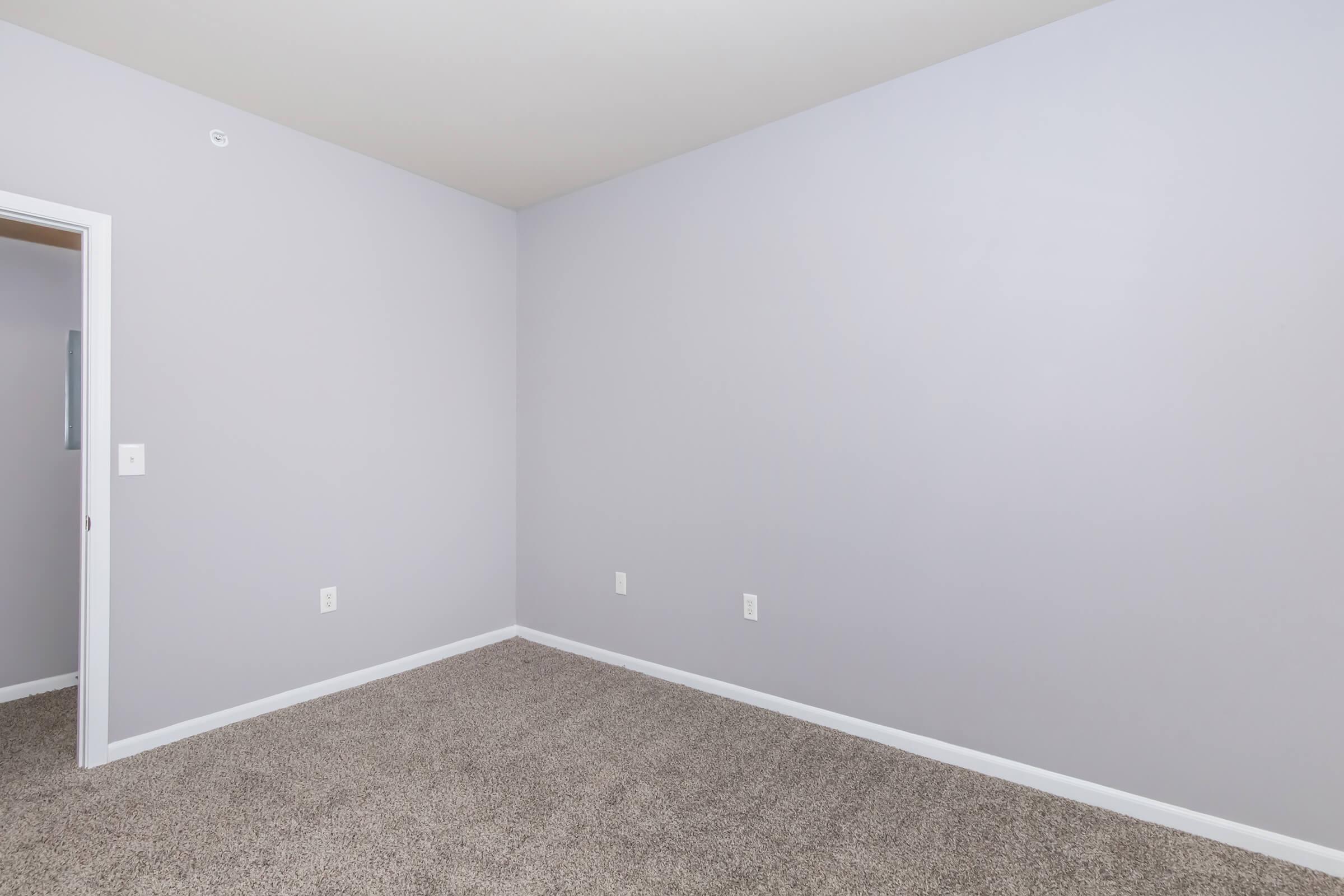 A spacious, empty room featuring light gray walls and a carpeted floor. The room has a single door on the left leading to an adjacent space. There are no furnishings or decorations, creating a blank canvas for potential use or decor.