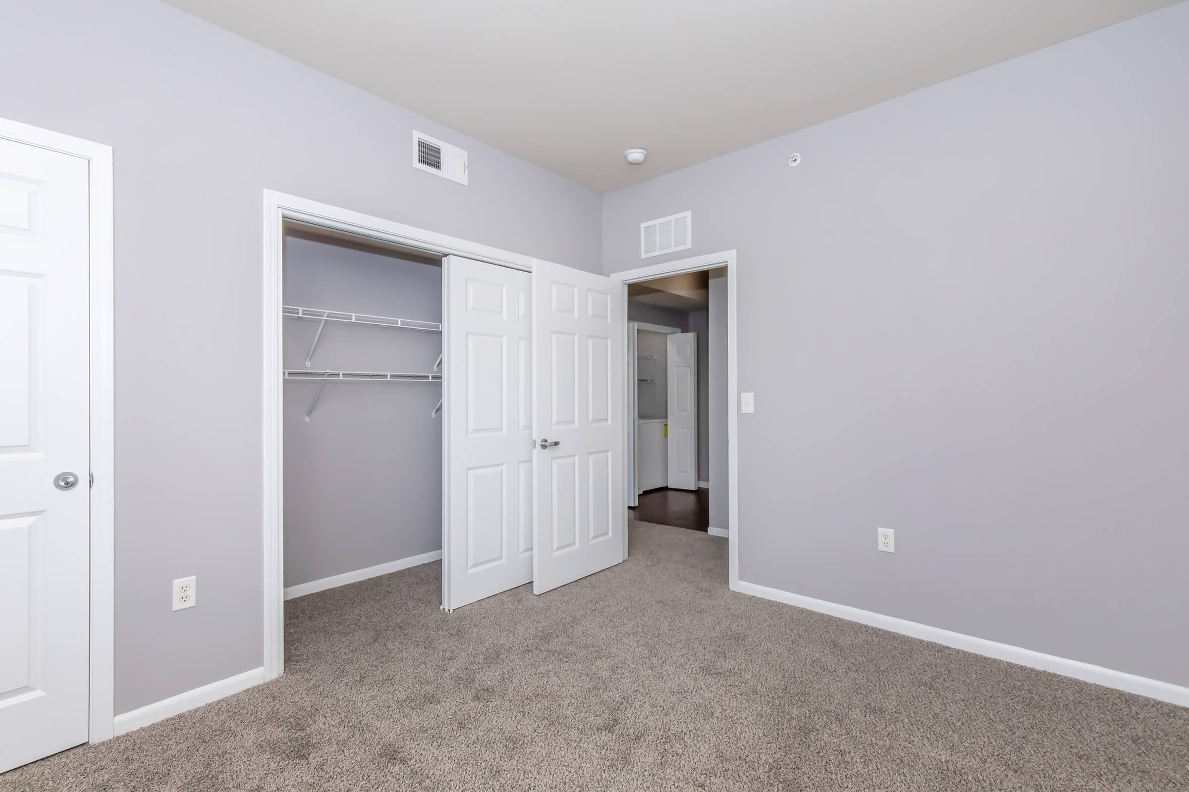 A well-lit empty bedroom featuring light gray walls and plush carpet. The room has two white double doors leading to an open closet with shelving. A doorway is visible to the right, leading to another room, while soft lighting adds warmth to the space.
