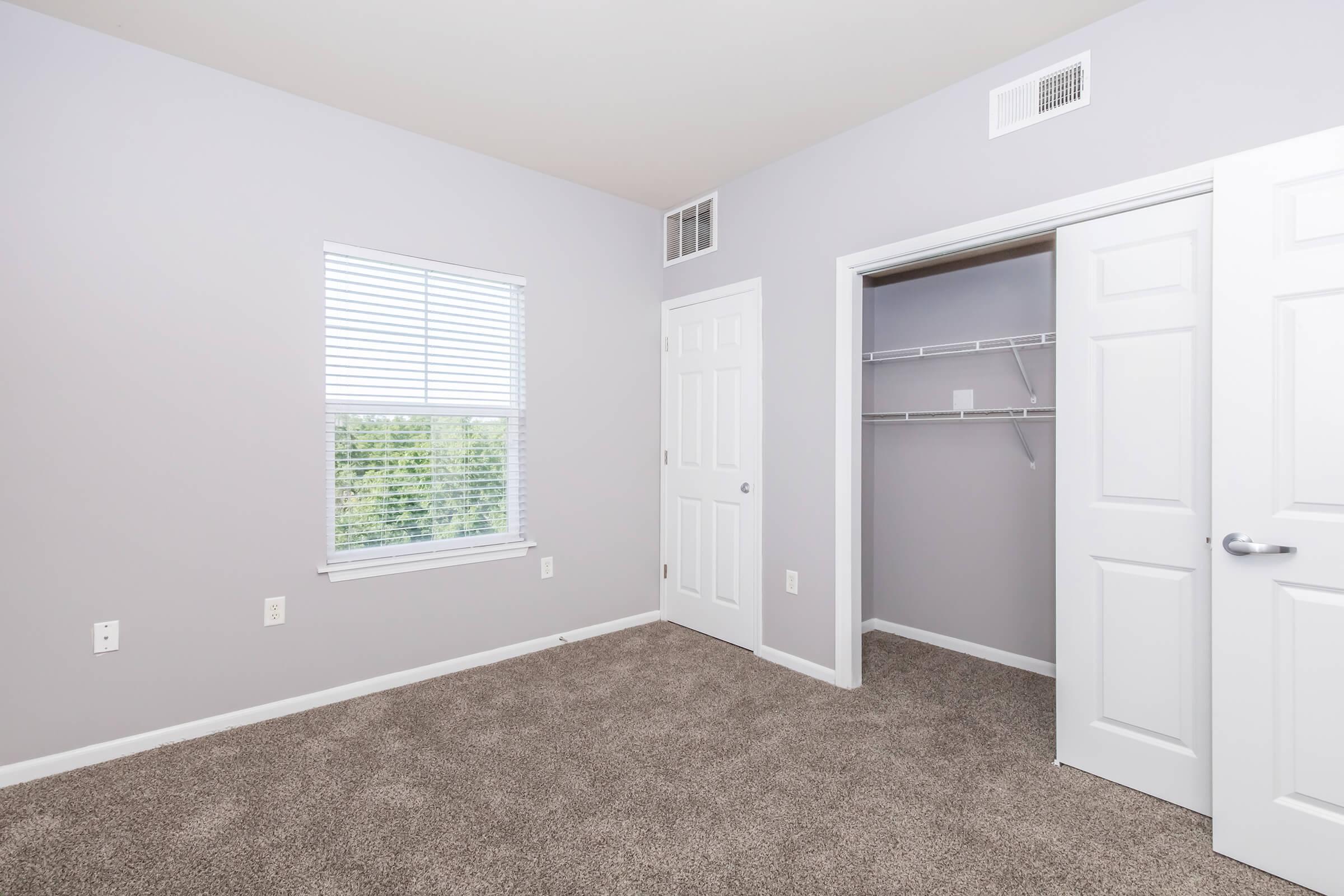 A spacious, well-lit room with light gray walls and carpet flooring. It features a window with white blinds, a closet with sliding doors, and a door leading to another area. The room is empty, providing a clean and inviting space.