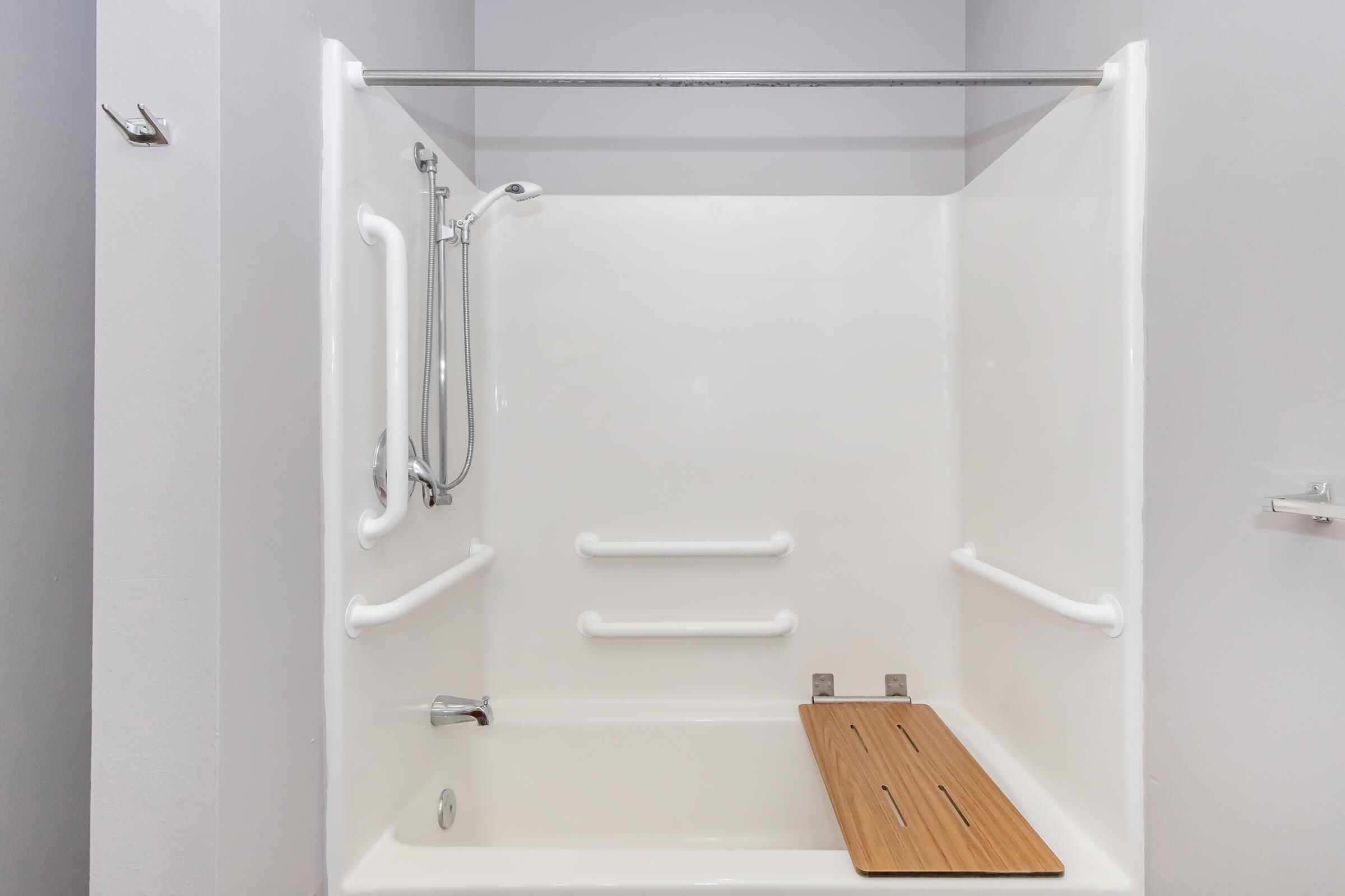 A clean, modern bathroom featuring a white shower stall with grab bars and a wooden shower mat. The walls are painted in a light gray color, enhancing the space's brightness and accessibility.