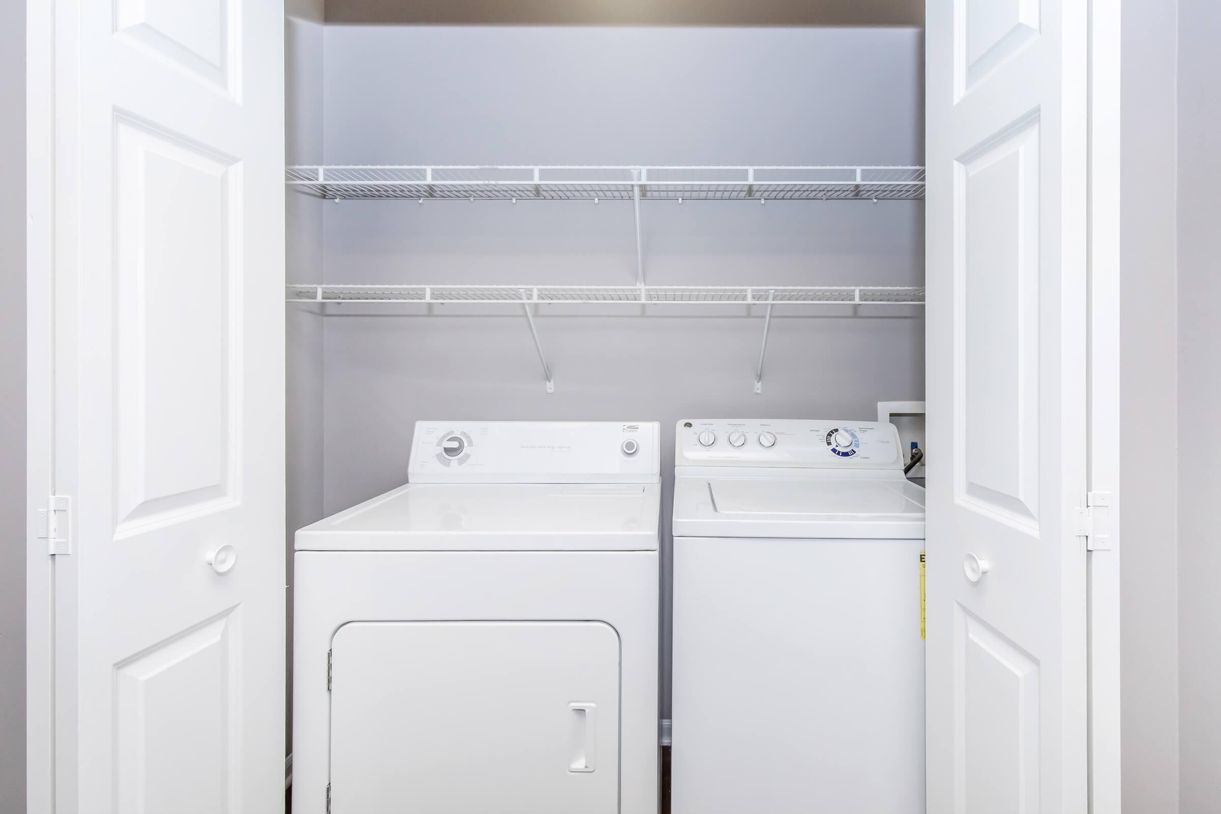 A laundry closet featuring a stacked washer and dryer, with white doors partially open. Above the appliances is a wire shelving unit for storage, against a neutral gray wall. The floor is not visible in the image.