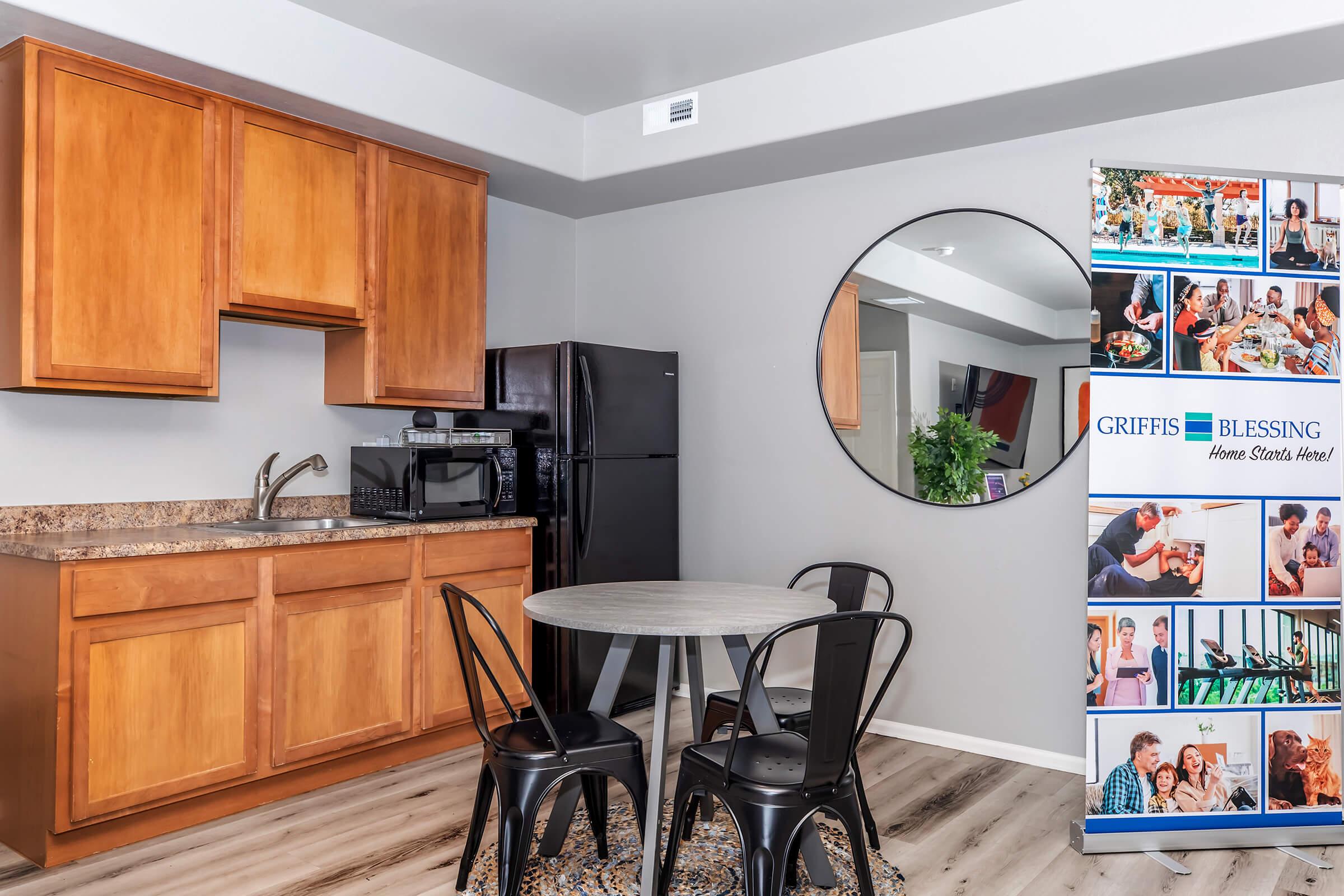 A modern kitchen with wooden cabinets, a black refrigerator, and a microwave, featuring a round table with three black chairs. A circular mirror hangs on the wall, and a promotional banner for Griffis Blessing is displayed nearby, showcasing images of people in communal settings.