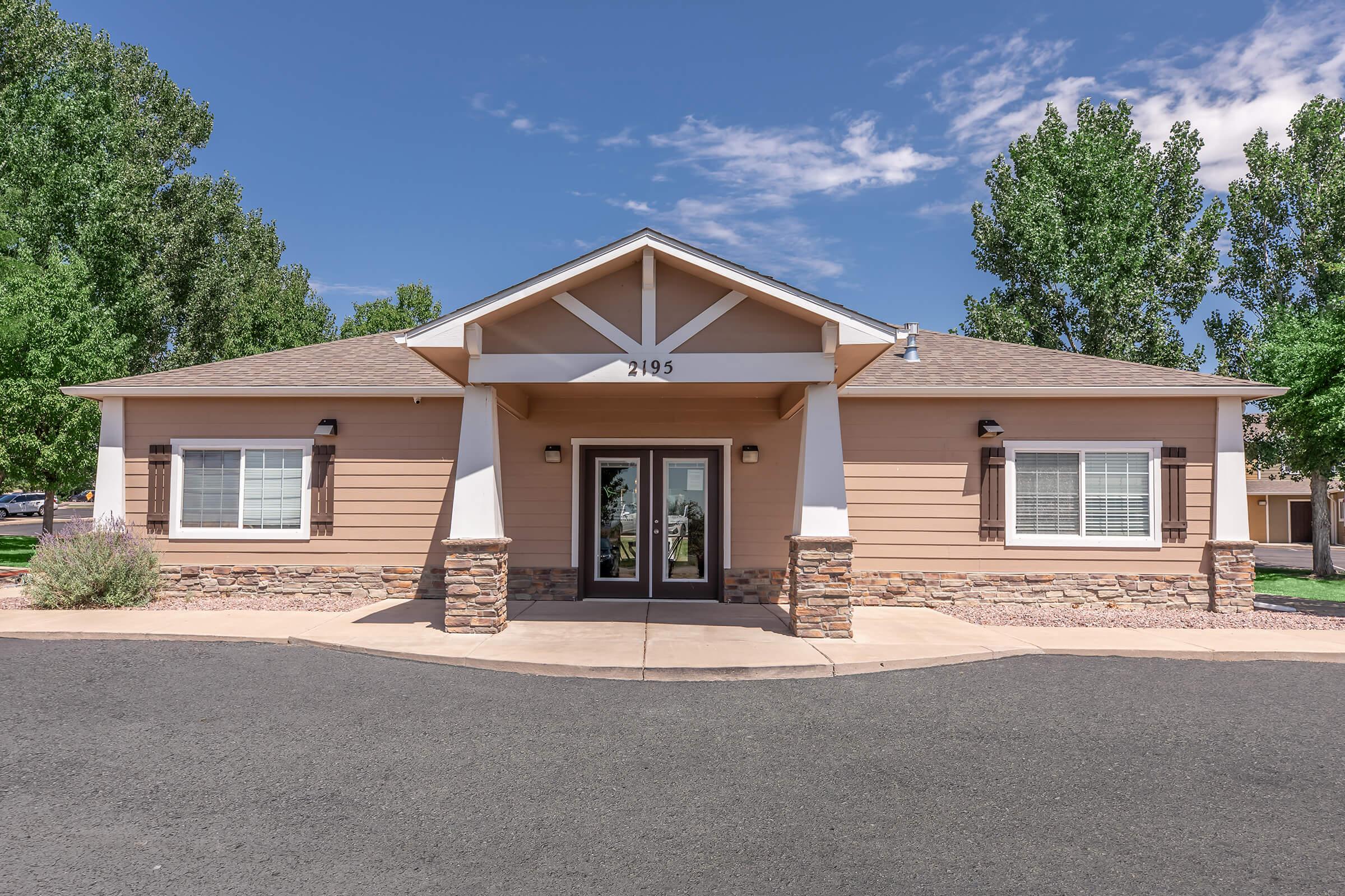 A single-story building with a light brown exterior and stone accents, featuring a gable roof. The entrance has a double door, flanked by windows on either side. The surrounding area includes green trees and a paved path leading up to the doorway. The sky is partly cloudy.
