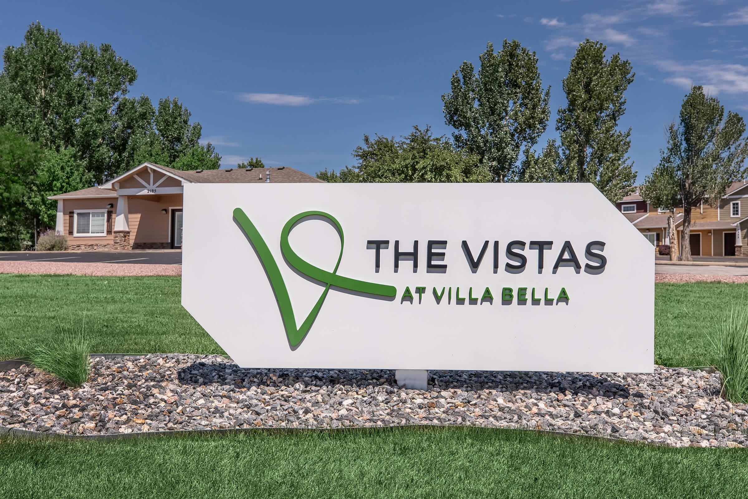 Sign for "The Vistas at Villa Bella" featuring a modern design with a green logo. The background includes well-maintained grass and trees, with residential buildings visible in a clear blue sky.