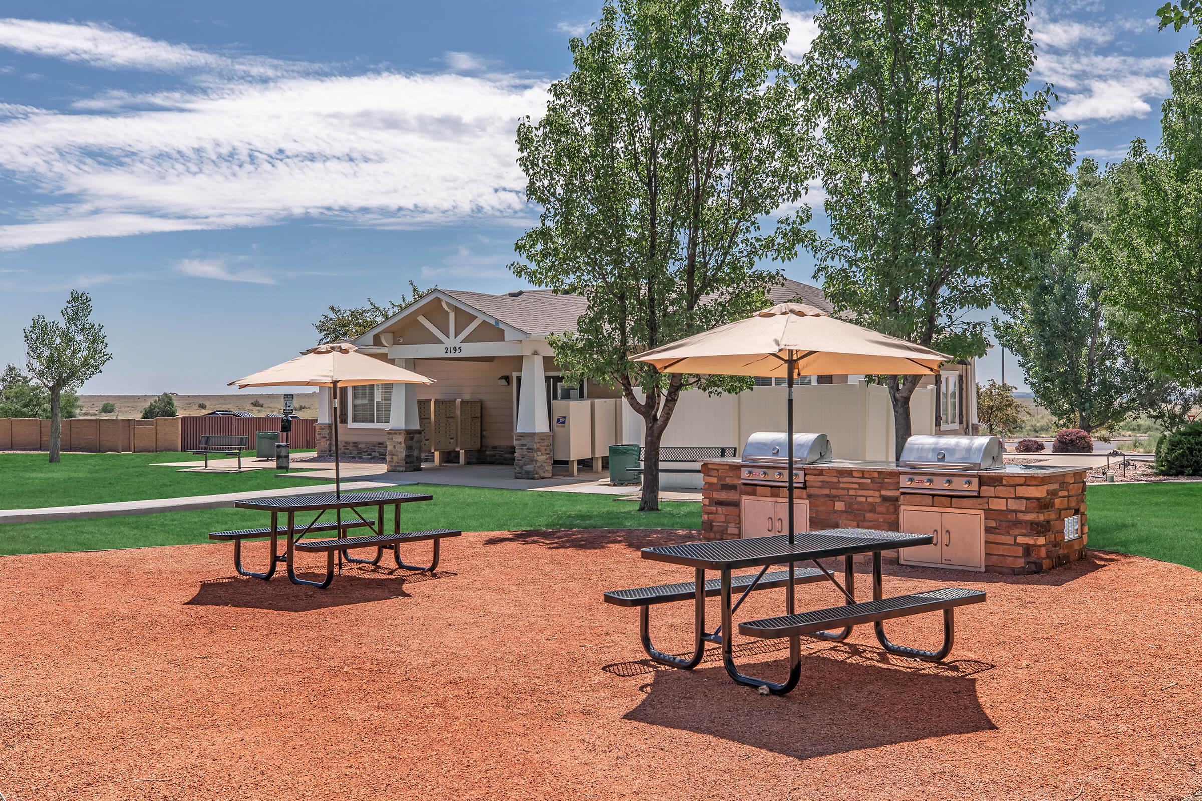 Outdoor picnic area featuring two black metal picnic tables with umbrellas on a landscaped turf. In the background, there is a building with a light-colored facade. Two silver grills are positioned nearby, surrounded by landscaped grounds with trees and a clear blue sky.