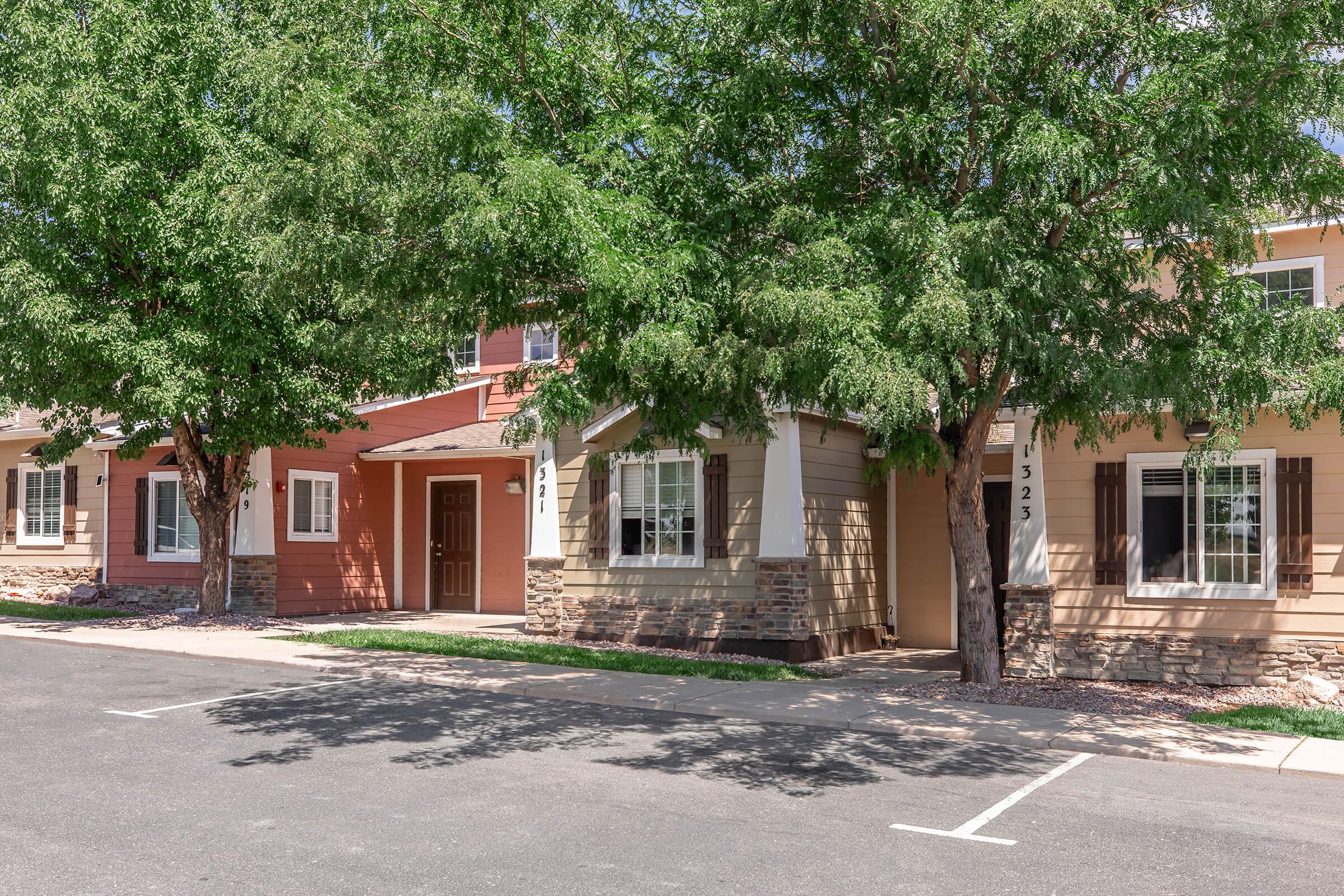 A row of modern houses in a suburban setting, featuring a mix of exterior colors including beige and red. Each house has large windows, and the front yards are shaded by leafy trees. A paved parking area is visible in front of the homes, with clear blue skies above.