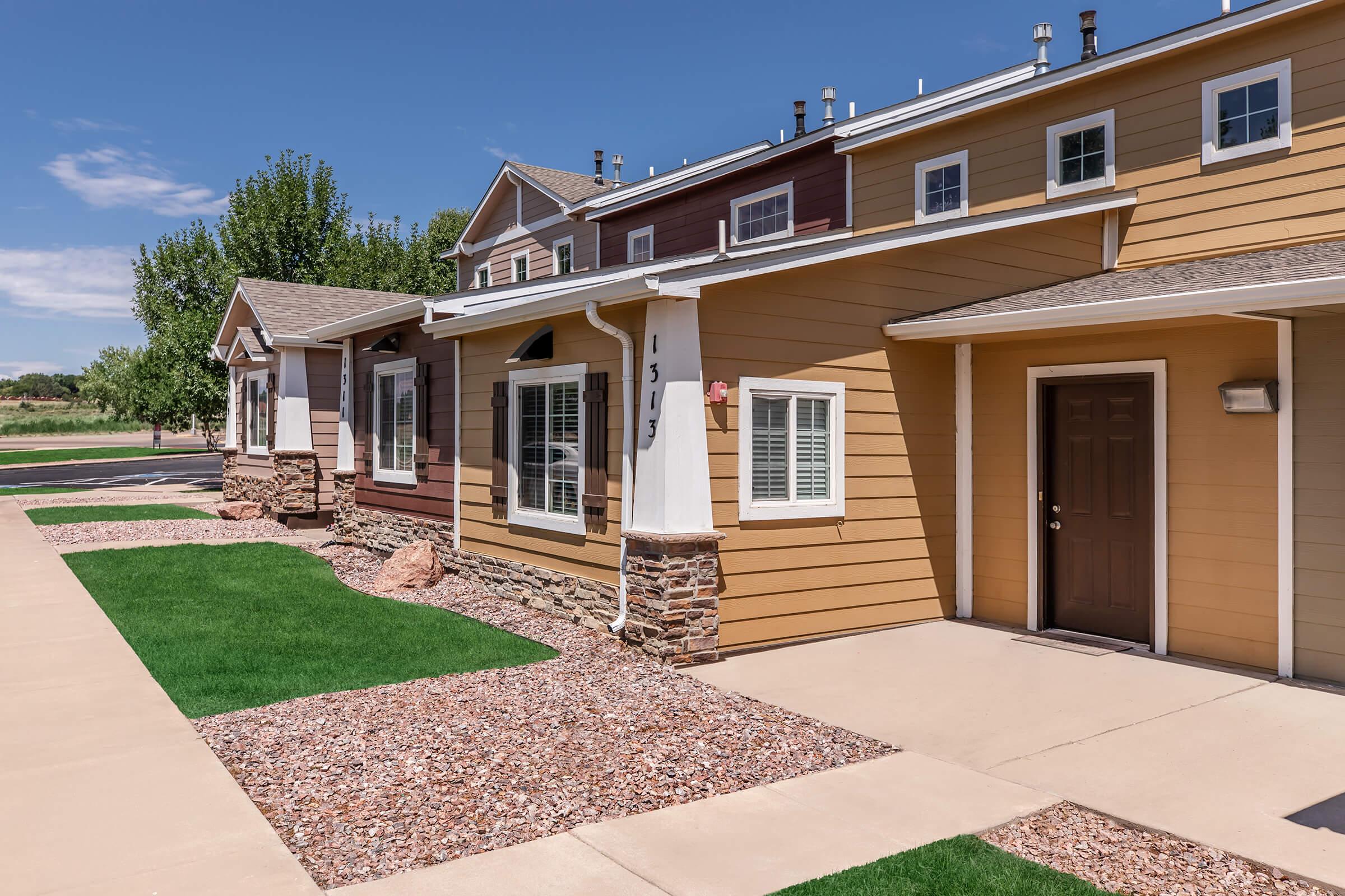 A well-maintained residential building with a combination of tan and brown siding, featuring neatly trimmed lawns and decorative stone accents. The property includes sidewalks and a clear blue sky, creating a welcoming atmosphere.