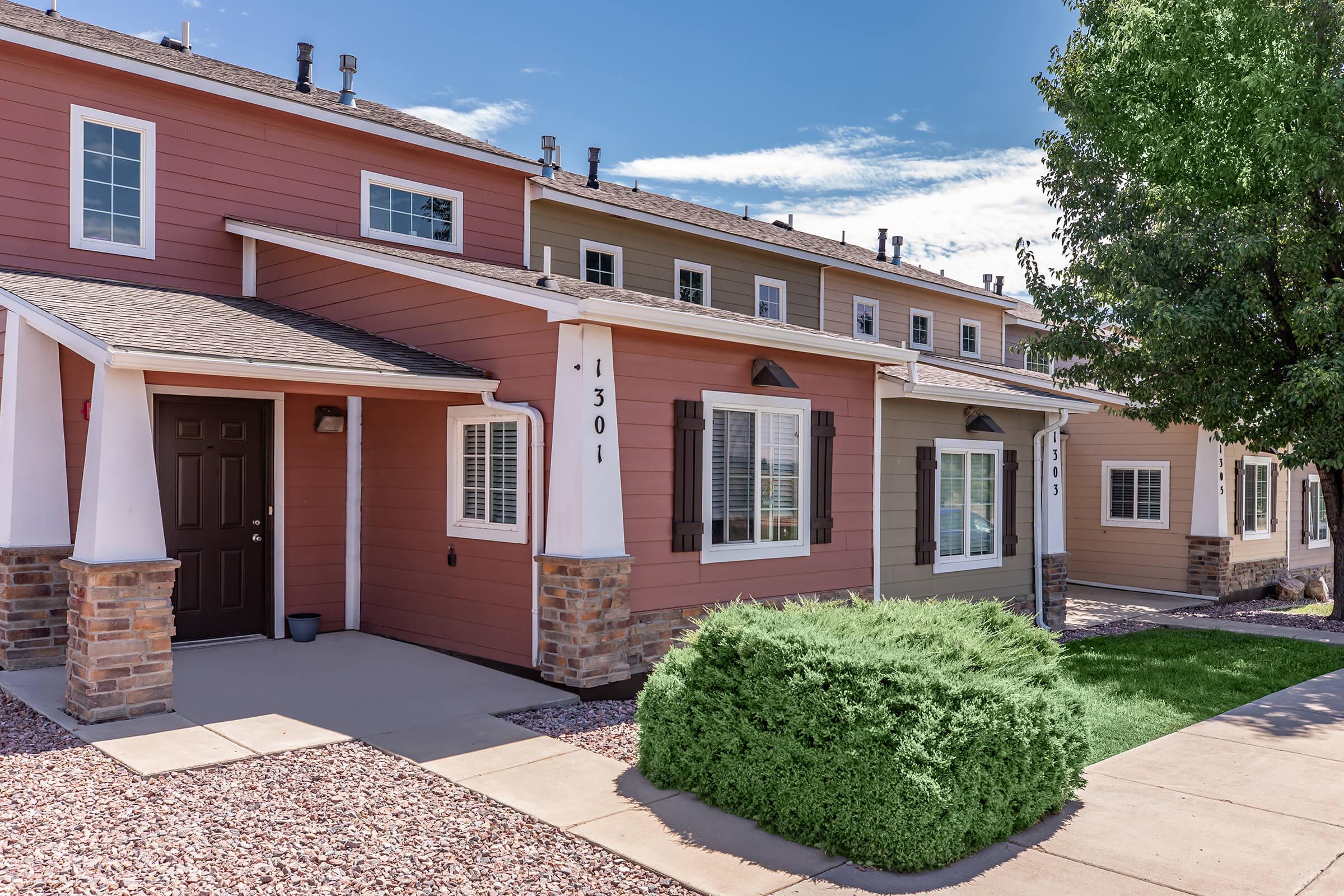 Row of two-story townhouses with colorful exterior siding in shades of pink and beige. Each unit features a small porch, windows with shutters, and landscaped yards with shrubs. A clear blue sky is visible above, creating a bright and inviting atmosphere.