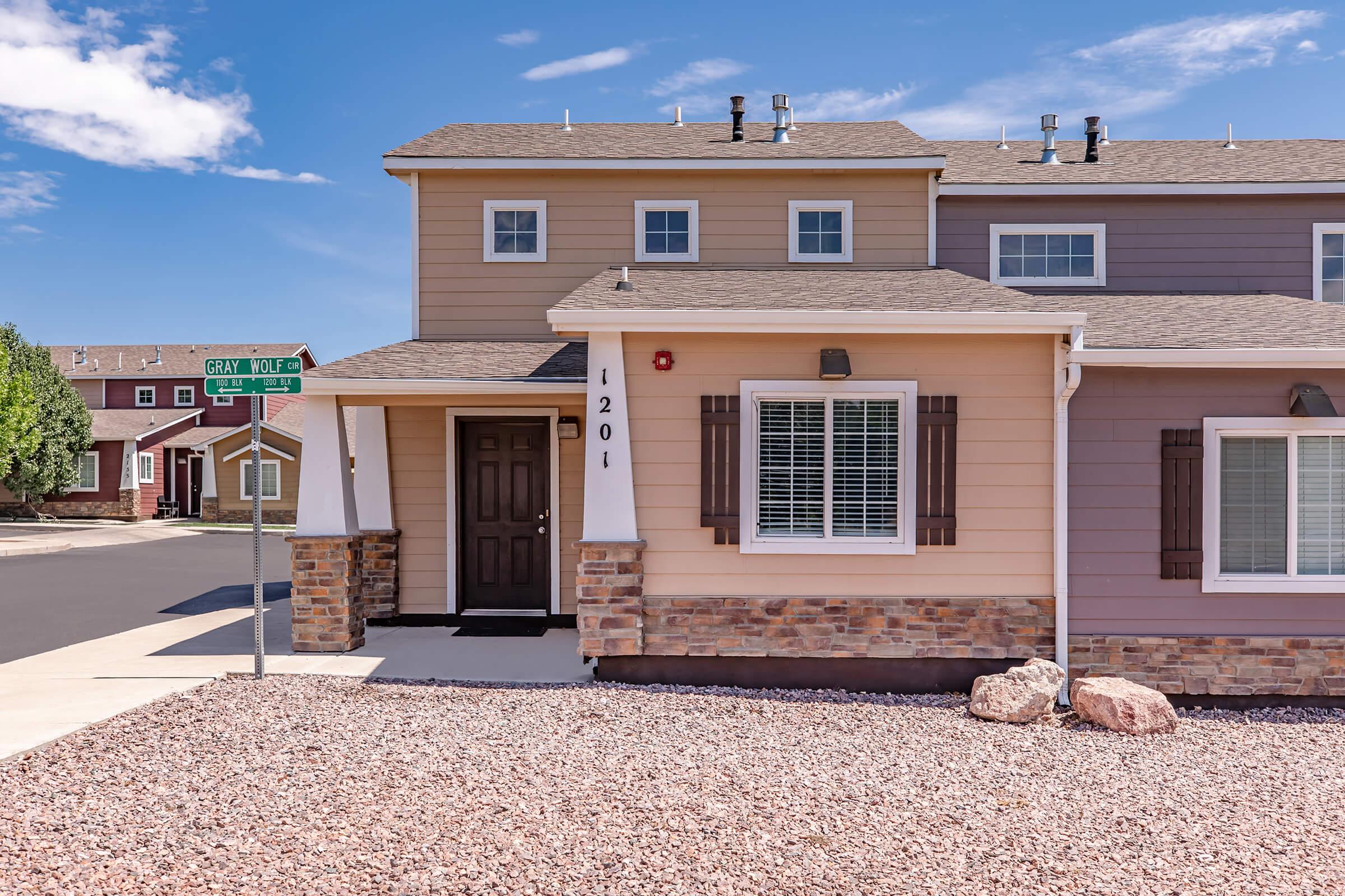 A two-story residential building with a stone foundation and light-colored siding. The entrance features a brown door and decorative shutters. Nearby, a street sign indicates "Gray Wolf" and "Eagle." The surrounding area has clear blue skies and is decorated with pebbles instead of grass.