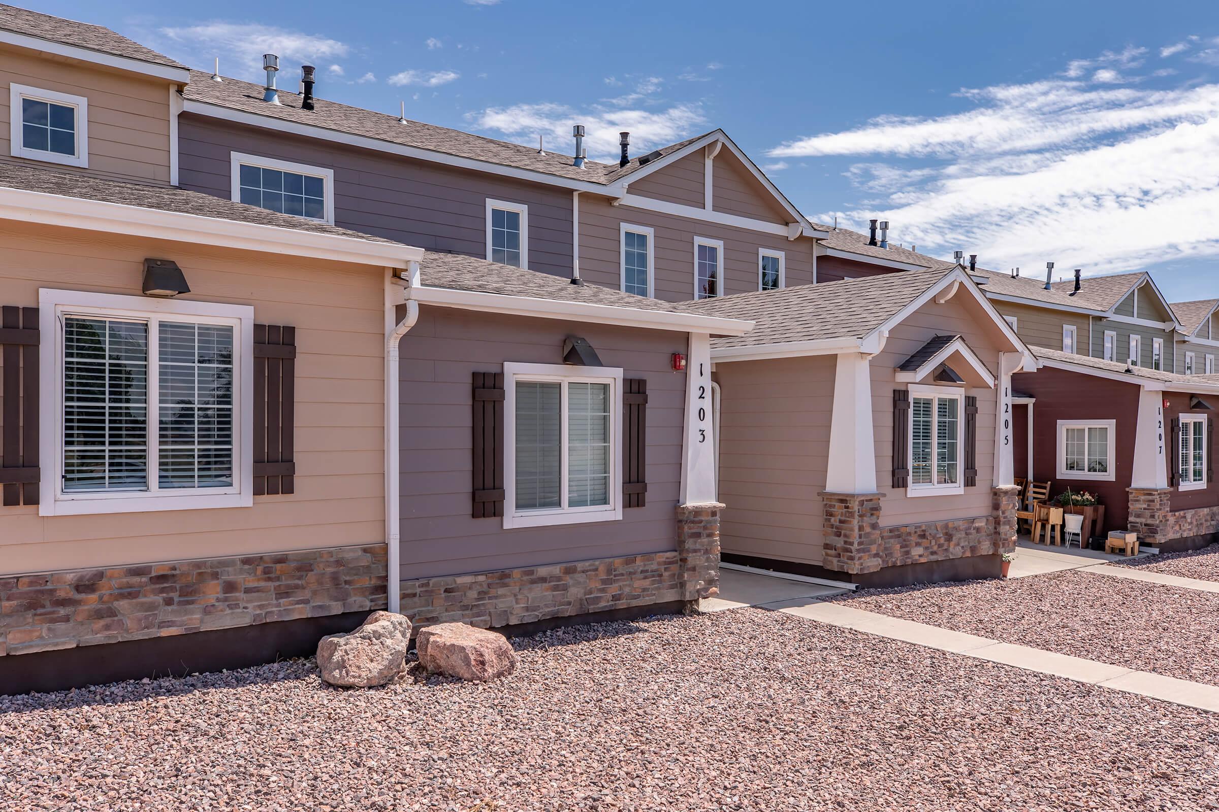 A row of modern townhouses with varied siding colors, featuring stone accents and large windows. The front yards have small rock landscapes and a sidewalk leading to the entrances. The sky is clear with a few clouds, suggesting a sunny day.