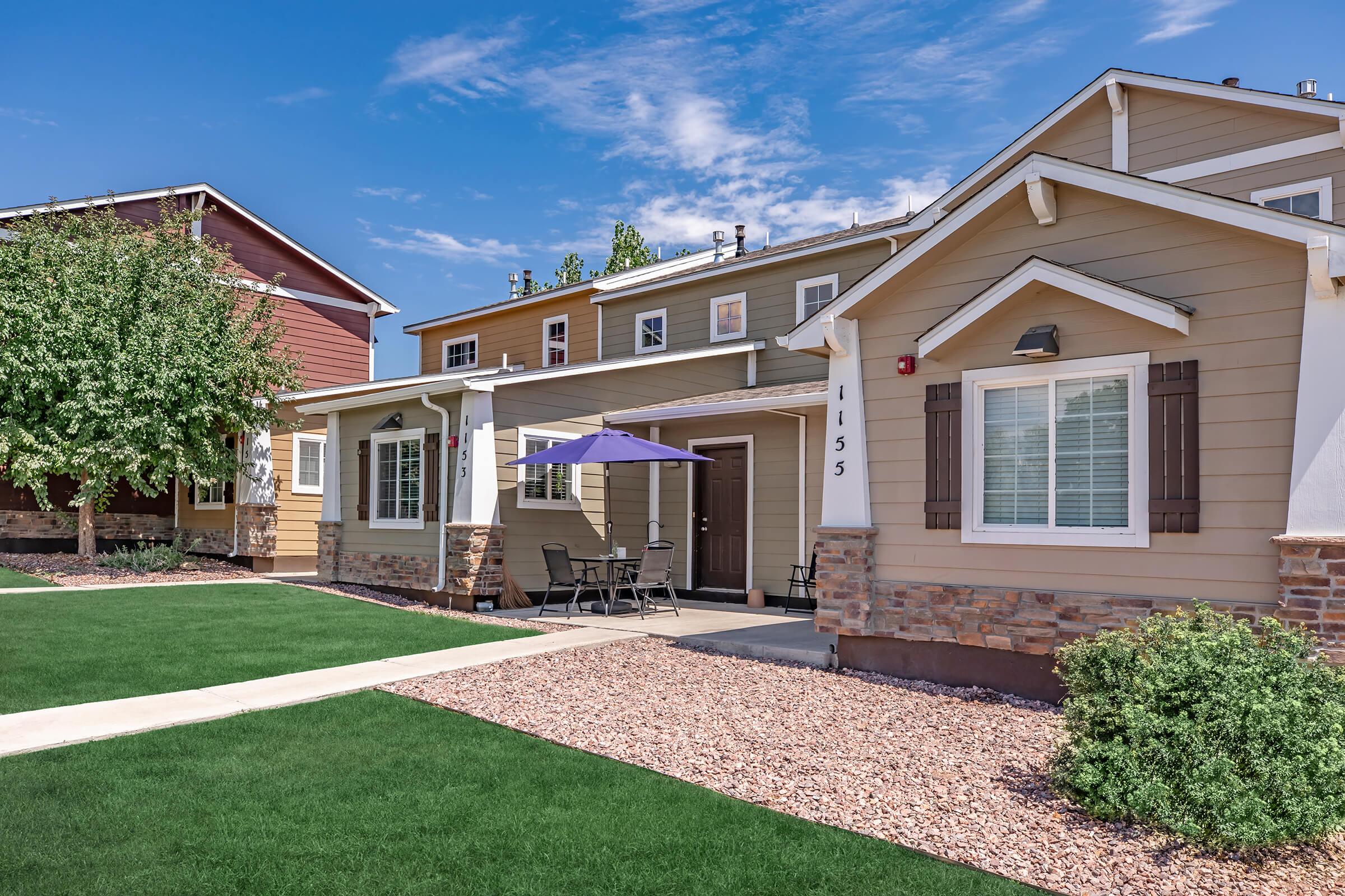 A well-maintained residential building with a light brown exterior and white trim. In front, there is a small patio area with a round table and chairs under a purple umbrella, surrounded by green grass and decorative gravel landscaping. Two trees are visible on the left side.