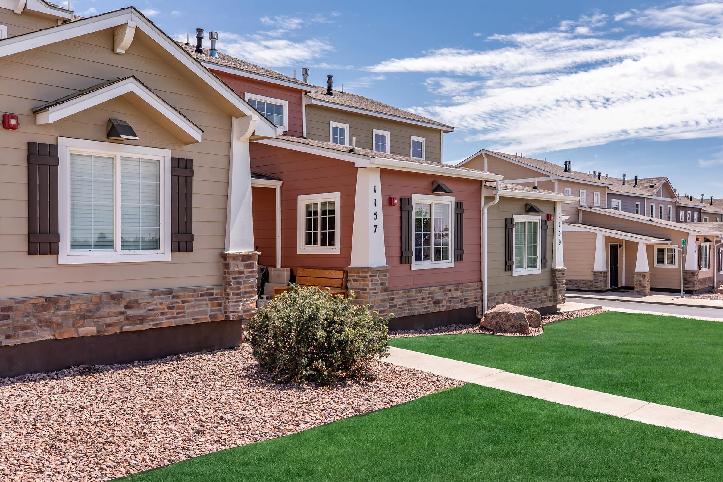 A row of modern homes featuring various exterior colors, stone accents, and manicured lawns. The scene includes a clear blue sky with scattered clouds, showcasing a well-maintained neighborhood with pathways and landscaping.