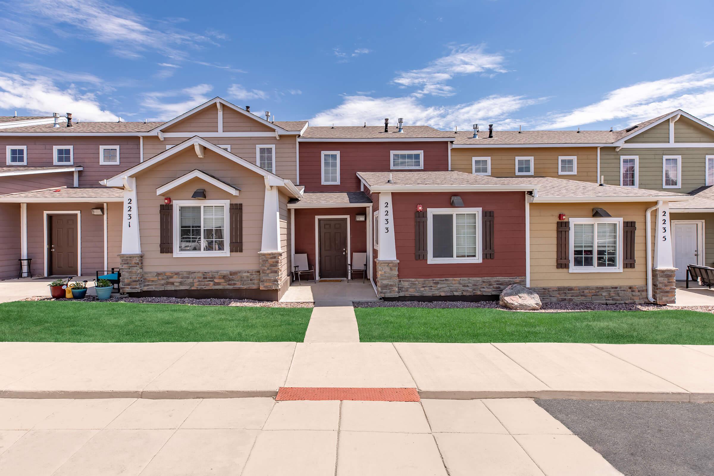 Two-story residential townhouses with colorful exteriors, featuring brick accents and shutters. Each unit has a small porch and front steps leading to the sidewalk. The vibrant houses are set against a clear blue sky with some clouds, and the well-maintained lawn adds to the appealing curbside view.
