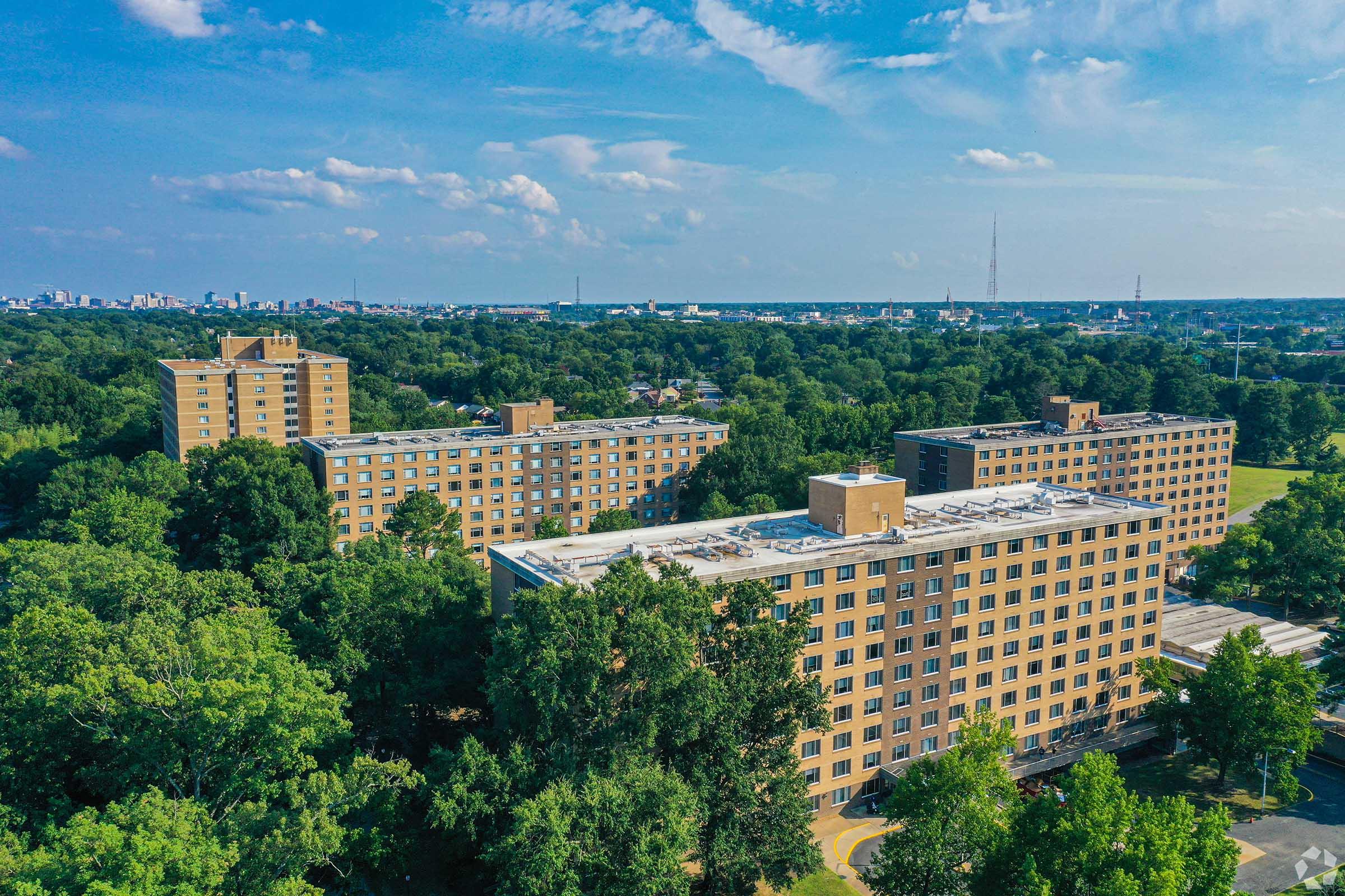 Aerial view of several large apartment buildings surrounded by green trees, with a city skyline visible in the distance under a partly cloudy sky. The scene conveys a mix of urban and natural environments.