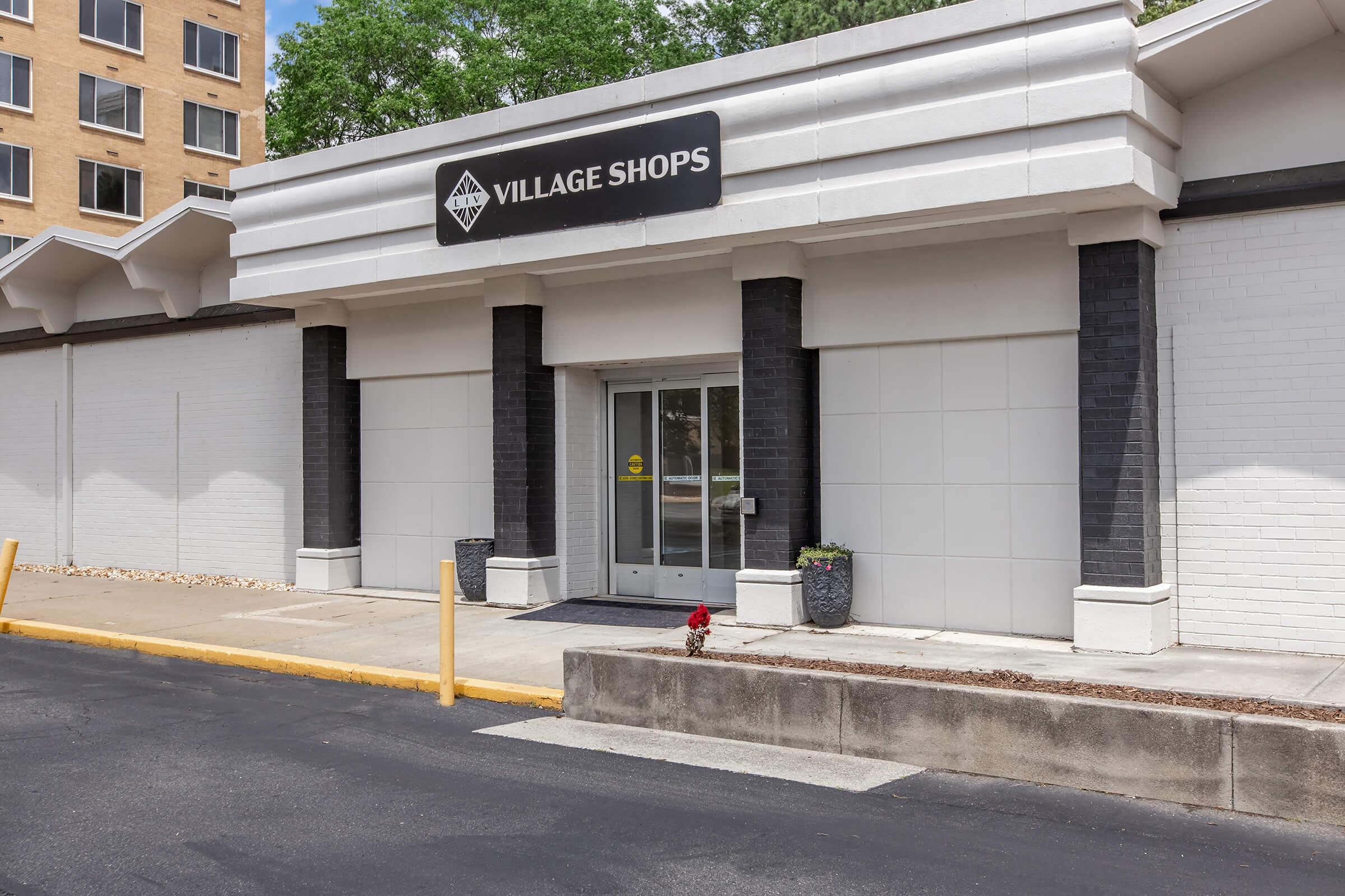 Exterior view of the Village Shops building, featuring an entrance with glass doors, flanked by black columns. The facade is mostly white, with a small flower pot on the side. The surrounding area includes a paved path and a low wall. A sign above reads “VILLAGE SHOPS.”