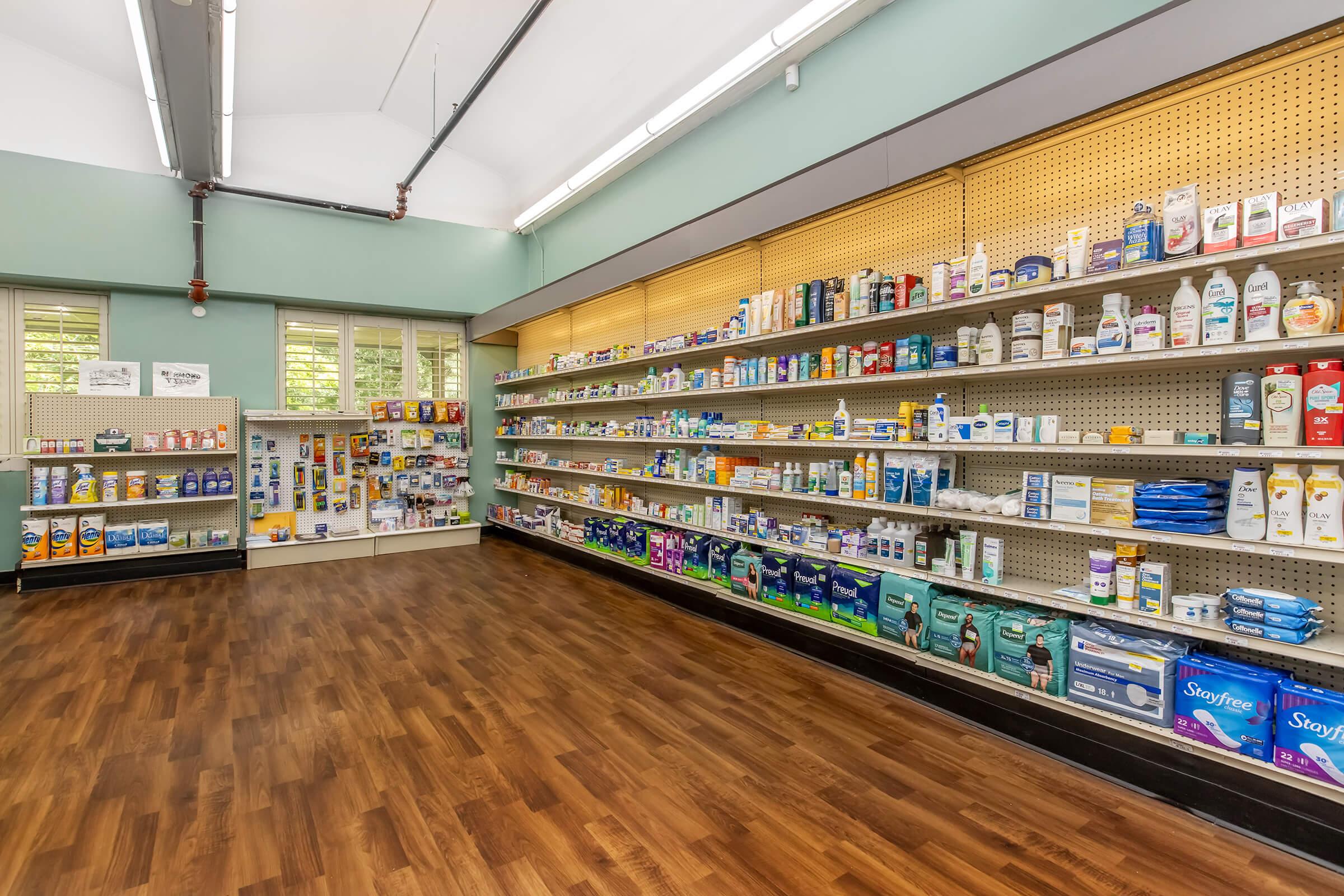 A well-organized aisle in a pharmacy or drugstore, featuring neatly arranged shelves stocked with various health and wellness products, including over-the-counter medications, personal care items, and toiletries, all set against a light blue wall and wooden flooring.
